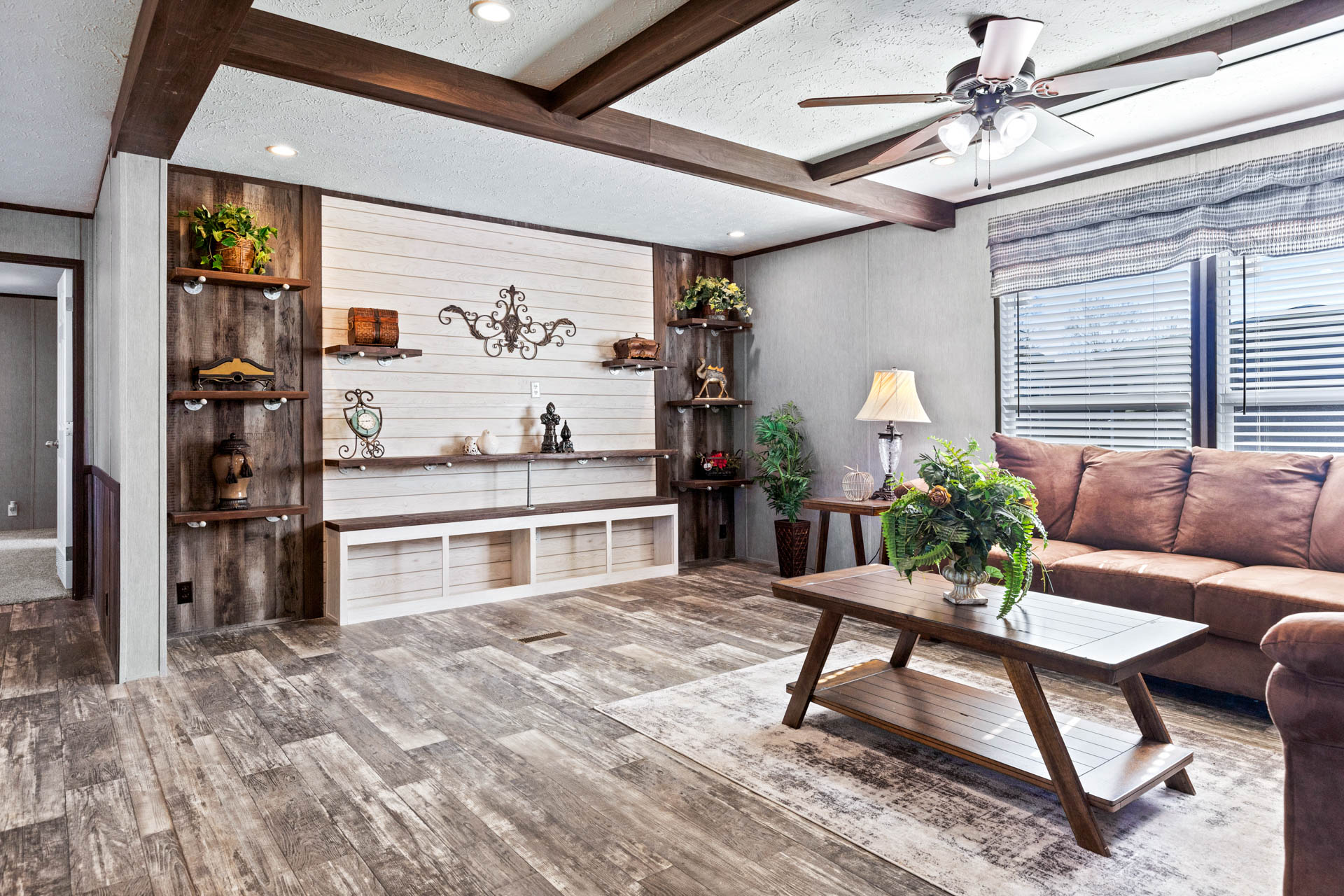 Cozy living room with rustic decor, featuring a brown sectional sofa, wooden coffee table with plant, shelves with ornaments, and large windows. Warm lighting.