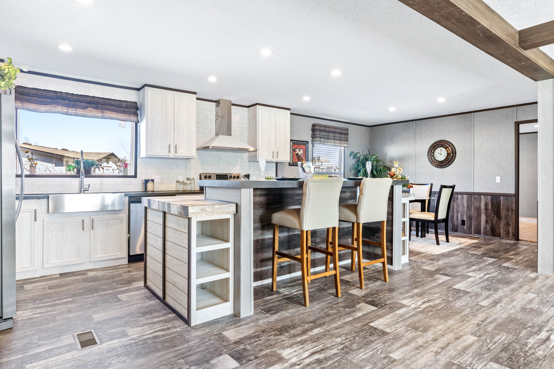 Modern kitchen with white cabinets, stainless steel appliances, and a large island with beige barstools. Dining area with clock on gray walls.