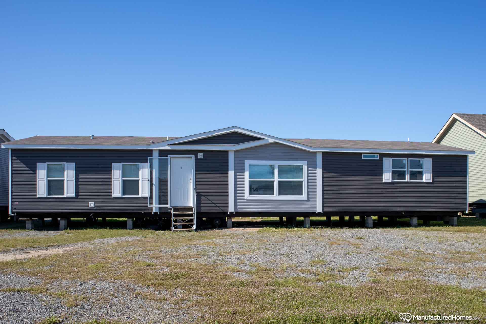 Brown, single-story manufactured home with white trim and large windows. It sits on a gravel lot, under a clear blue sky, conveying simplicity and openness.