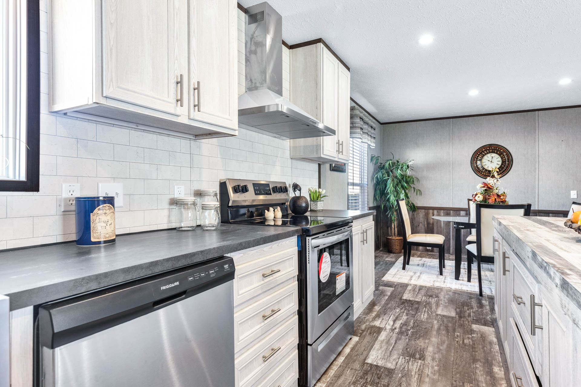 Modern kitchen with stainless steel appliances, white cabinets, and dark countertops. A dining area with a round table, floral arrangement, and wall clock is visible. The space feels bright and inviting.