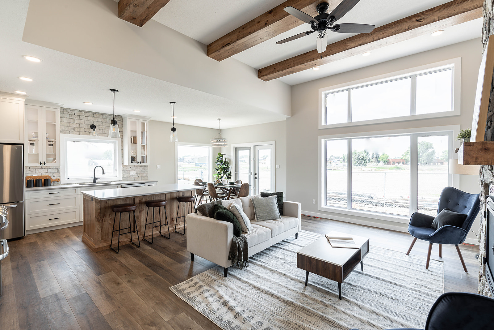 Modern open-concept living room and kitchen with wooden beams, large windows, a beige sofa, blue armchair, and a dining area, conveying a cozy ambiance.