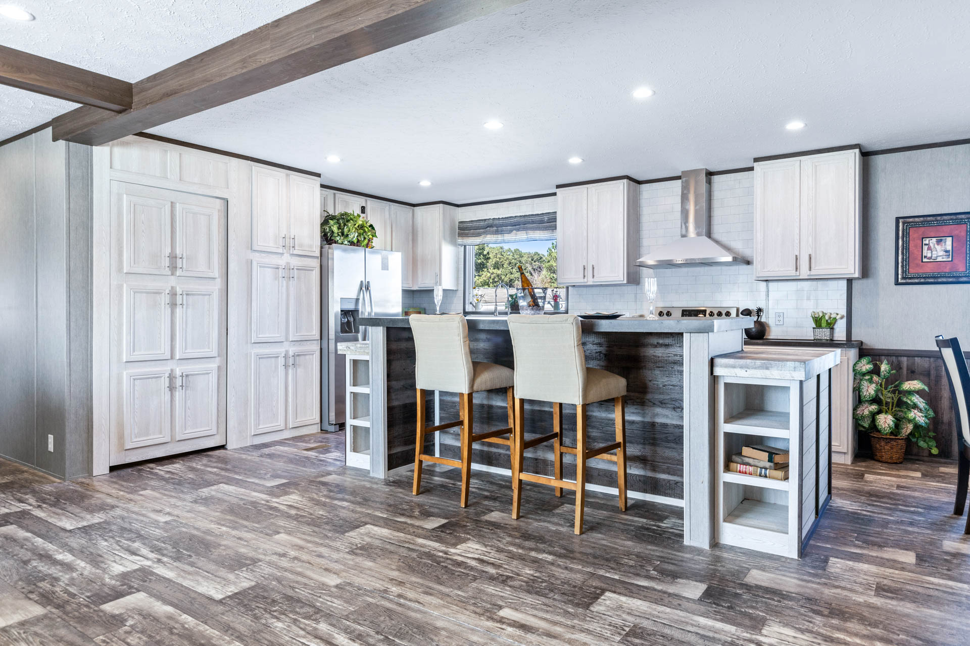 Modern kitchen with white cabinets, stainless steel appliances, and wood accents. Two beige barstools at a dark wood island. Natural light fills the space.