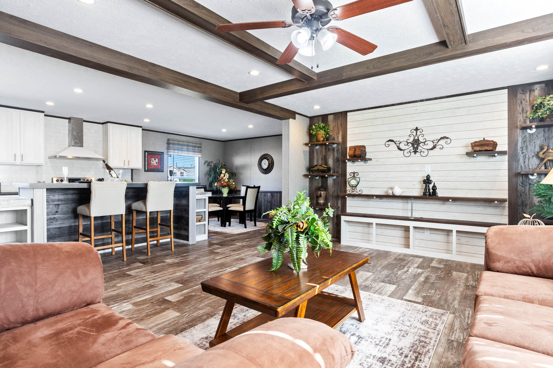 Modern open-plan living room with wooden beams, brown sofas, and a ceiling fan. Features a cozy kitchen with bar stools and a dining area.