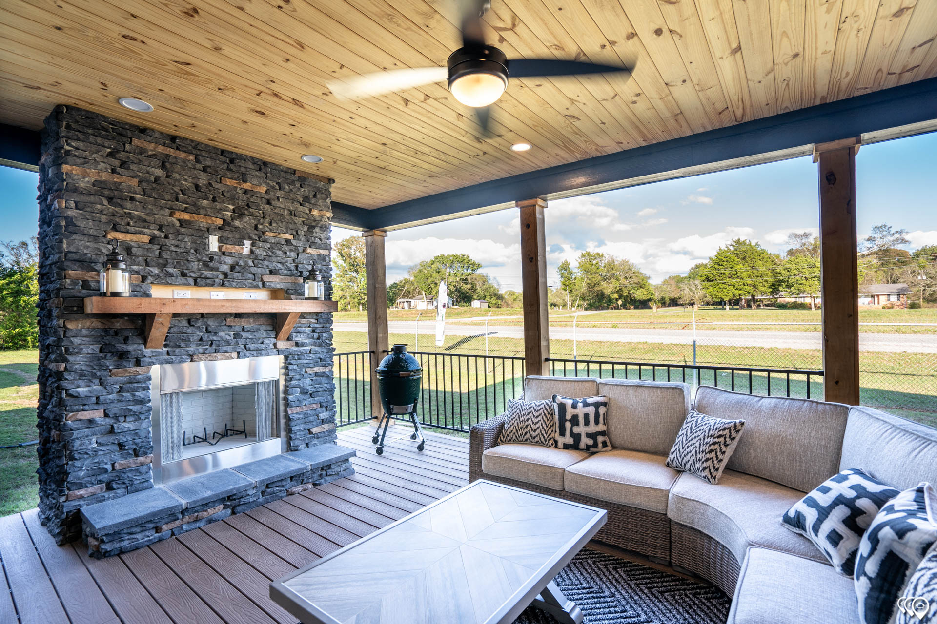 Covered outdoor patio with stone fireplace and comfortable seating. Ceiling fan above, grill nearby, overlooking a sunny, tree-lined lawn. Cozy and inviting.