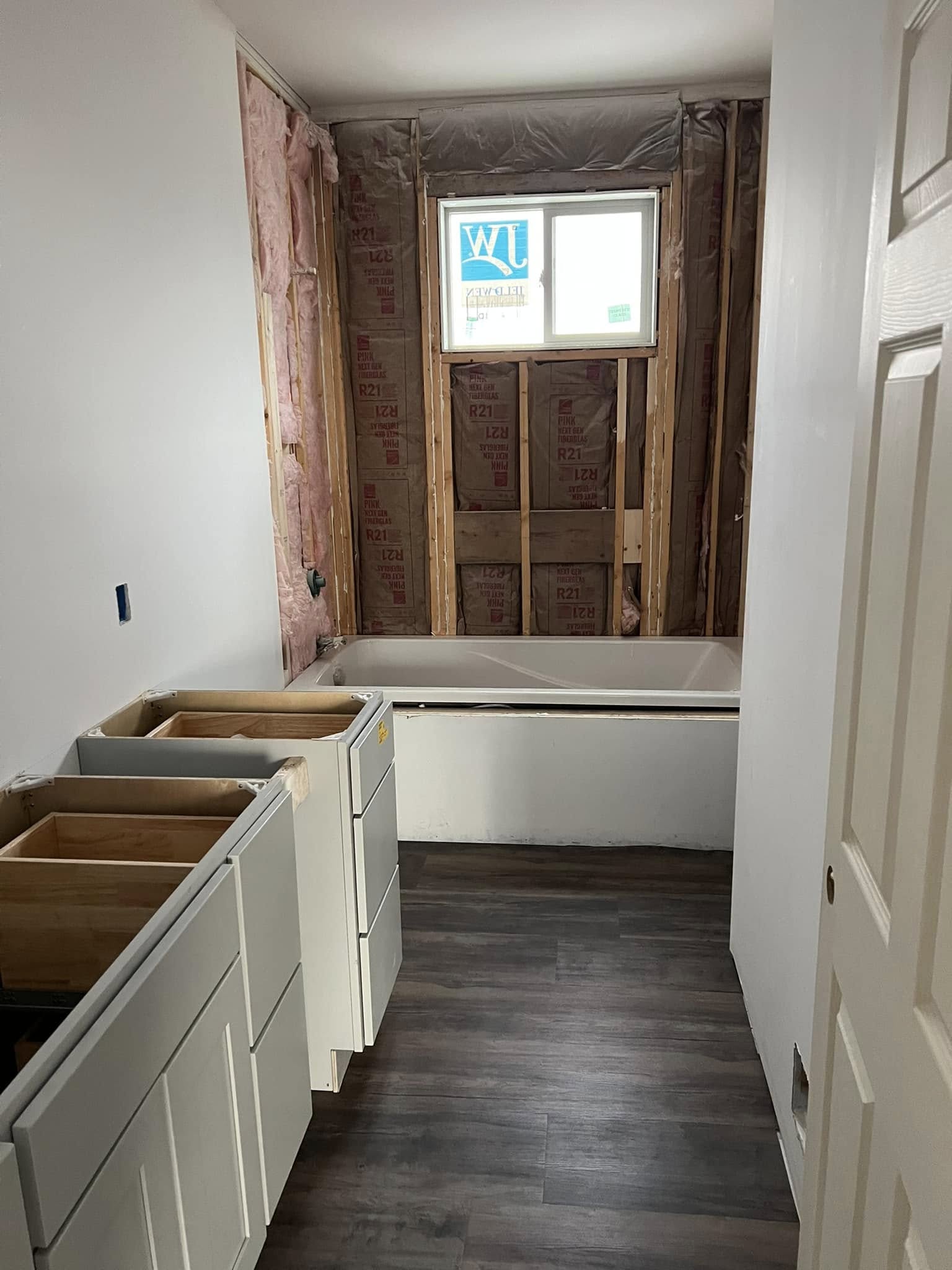 Partially renovated bathroom with exposed insulation, wood framing, and a small window. Unfinished cabinets are installed on the left, with a bathtub at the far end. The space has a work-in-progress feel.