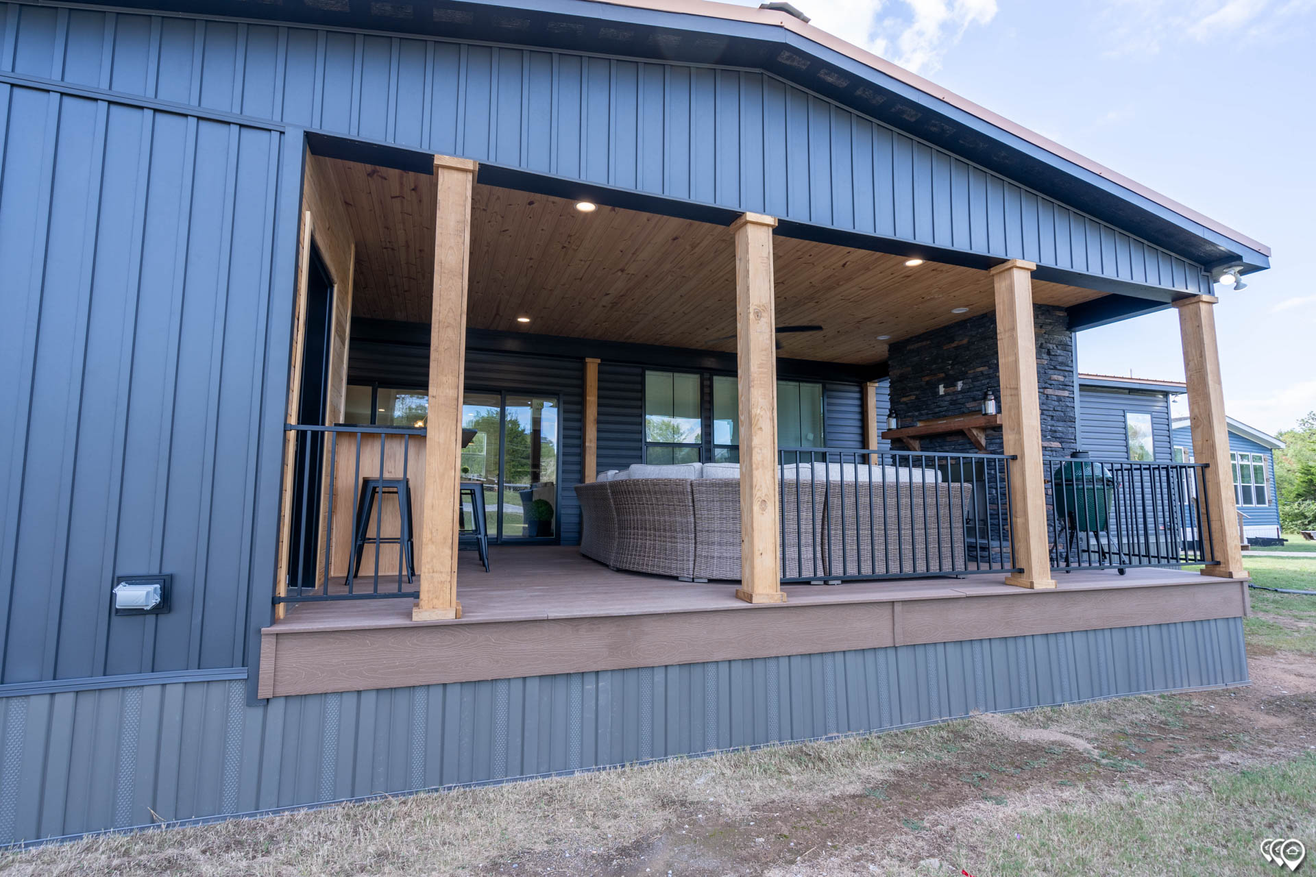 Covered wooden deck with light wood beams and dark siding on a modern house. Features cozy seating, a stone fireplace, and tranquil outdoor views.