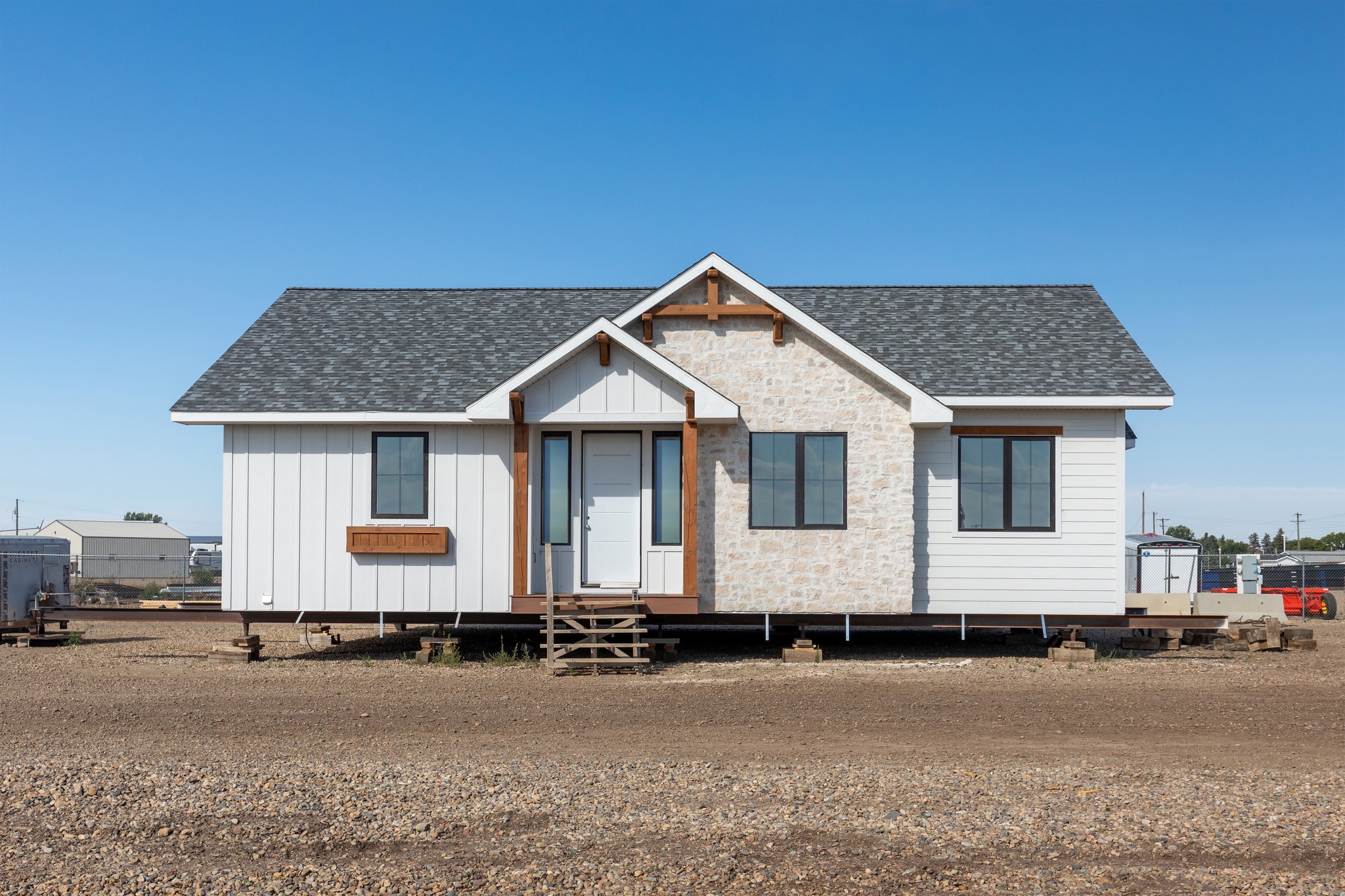 Single-story, modular home on raised blocks, featuring white siding, stone accents, and a dark roof. Clear blue sky backdrop enhances the serene setting.