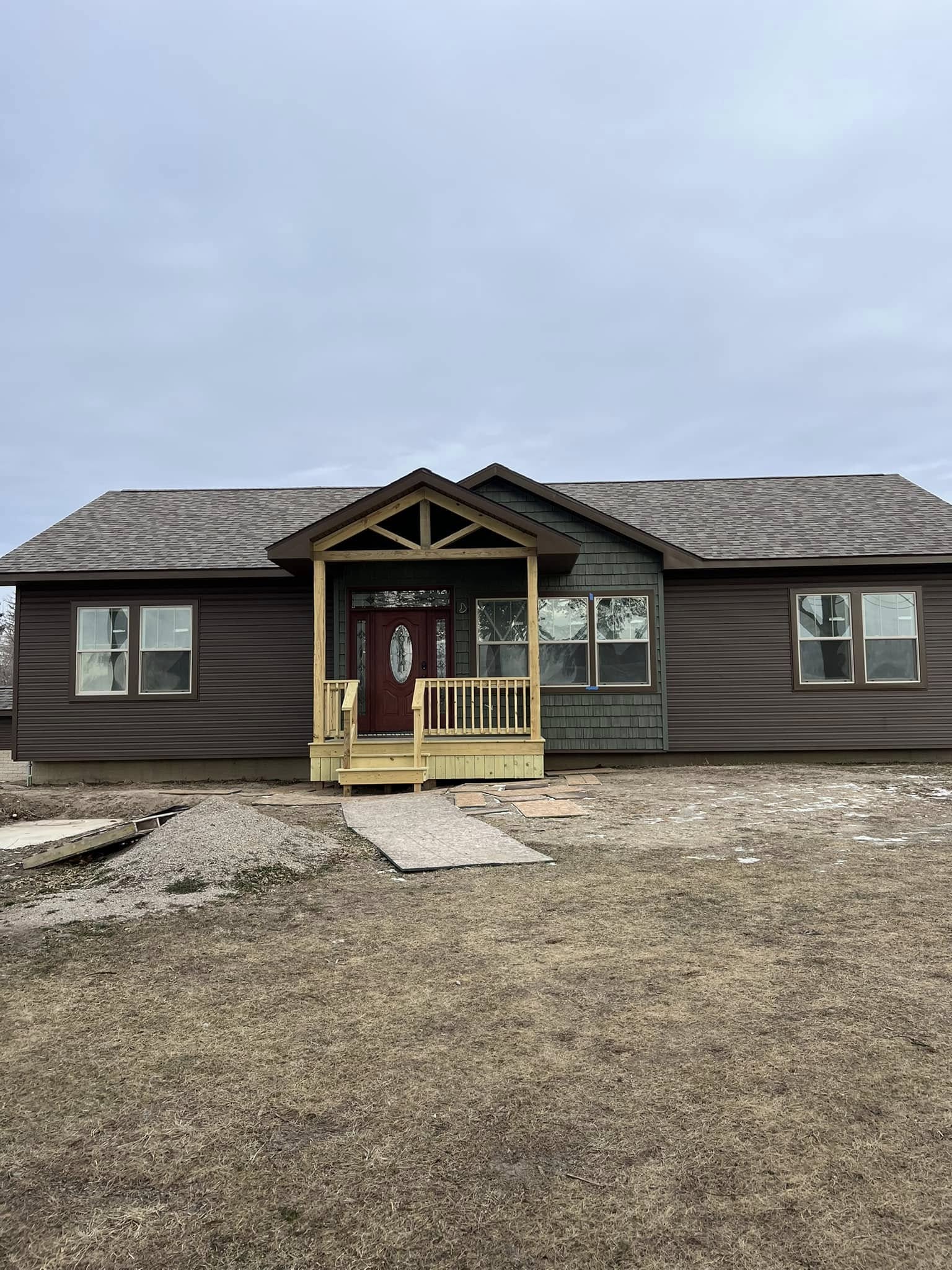 A single-story house with brown siding and a pitched roof. It features a small front porch with a red door and decorative wooden beams. The yard is bare with patches of grass. The sky is overcast, creating a calm atmosphere.