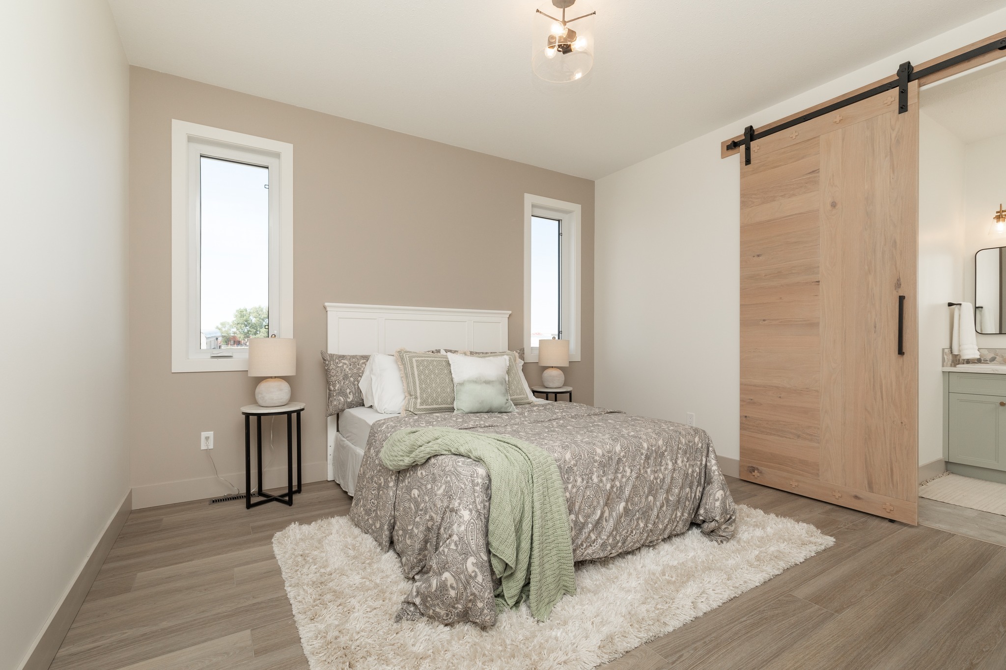 Minimalist bedroom with a cozy vibe features a bed with gray bedding and green accents. Two windows frame nightstands with lamps. A wooden sliding door leads to a bathroom. Light tones and simple decor create a serene atmosphere.