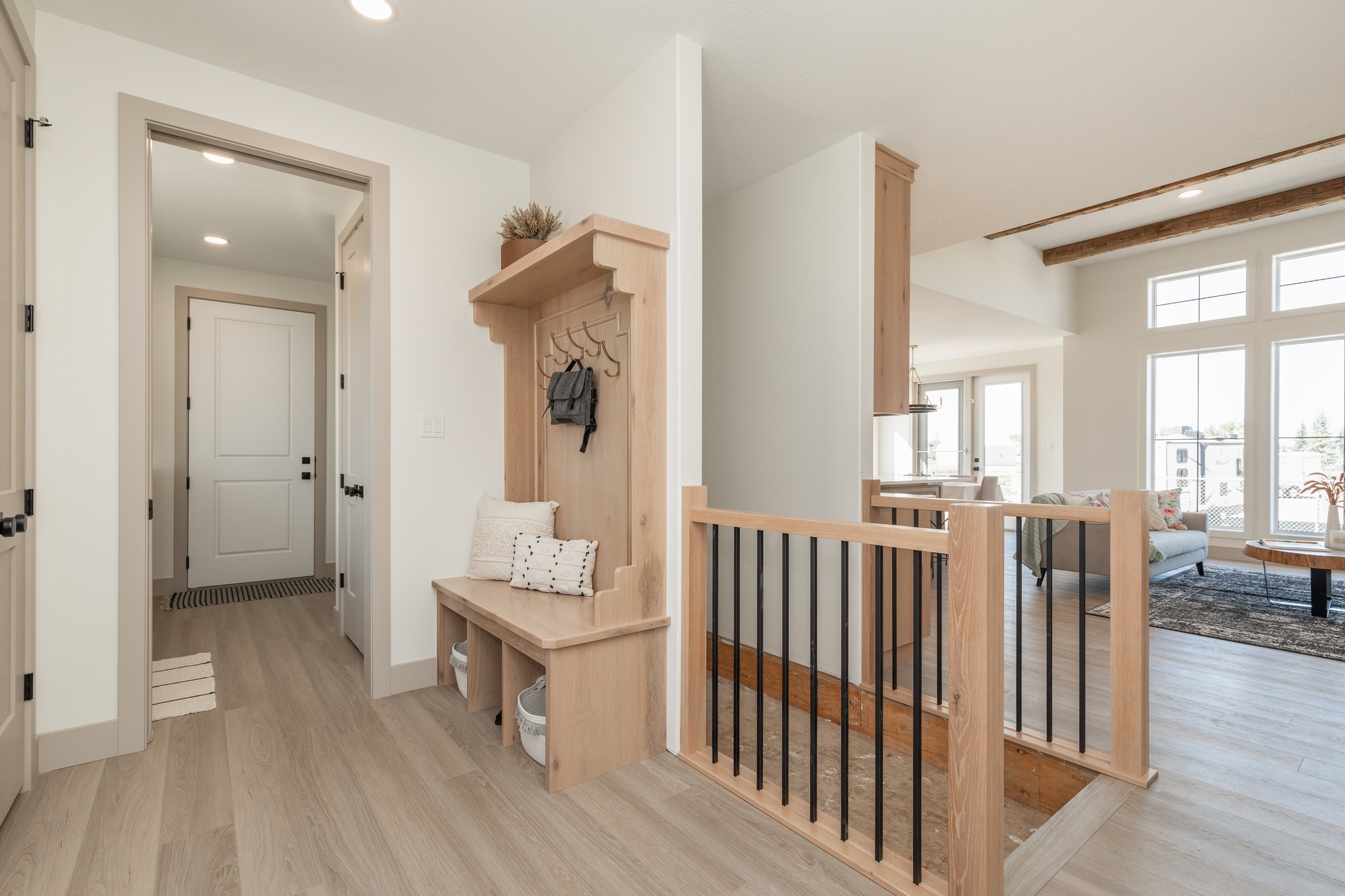 Bright, modern entryway with light wood floors and a cozy bench with hooks. Open staircase leads to living room with large windows and natural light.