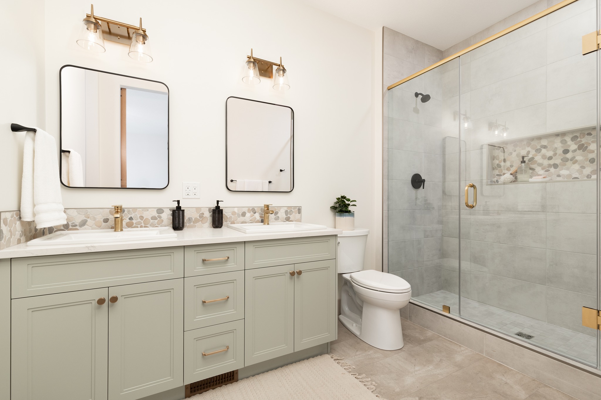 Modern bathroom with gray vanity, dual sinks, and black fixtures under square mirrors. Glass shower with gold trim, tiled walls, and white toilet.