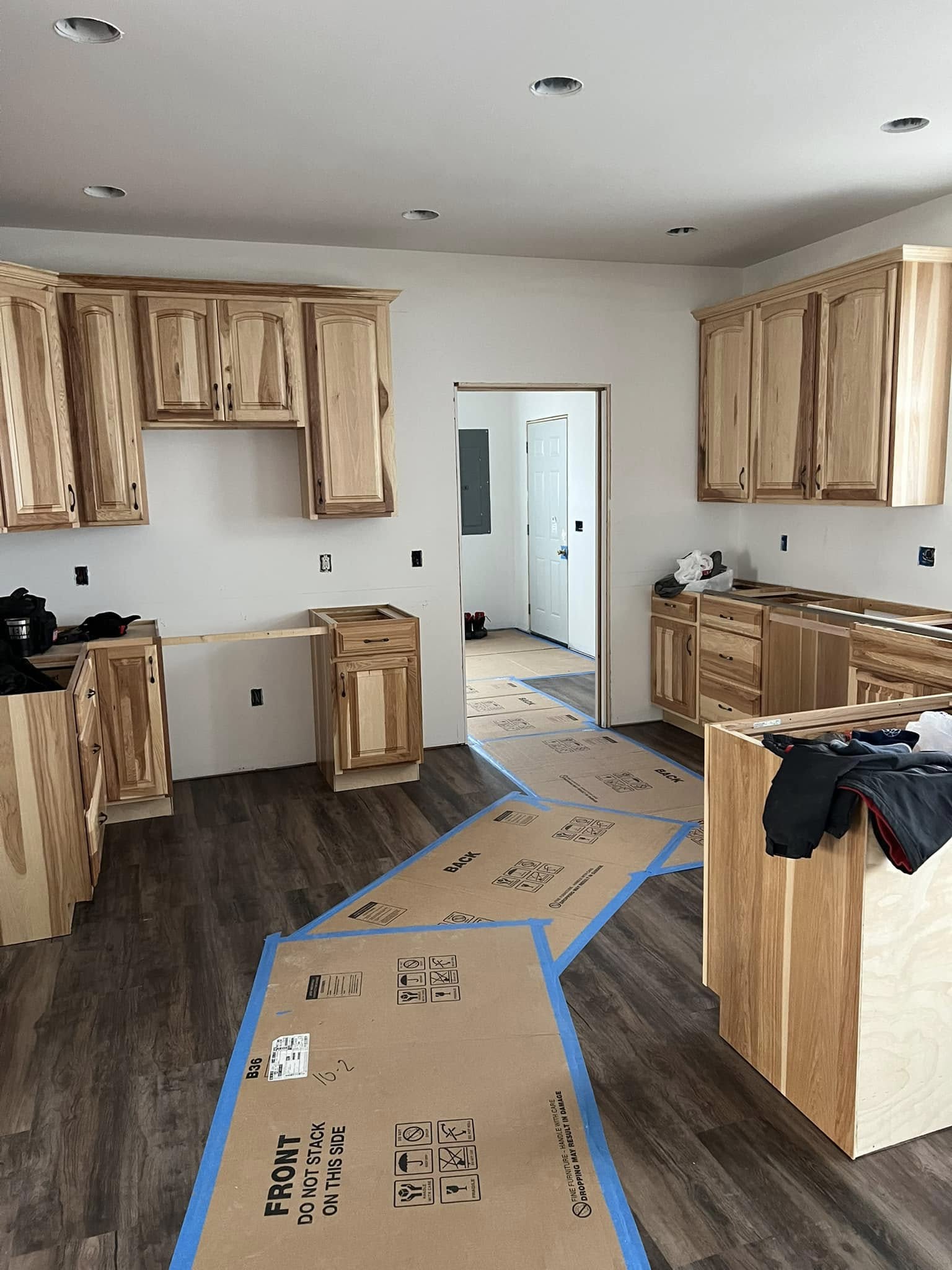 A kitchen under construction with light wood cabinets and dark flooring. Cardboard covers the floor, suggesting ongoing work. The tone is industrious.