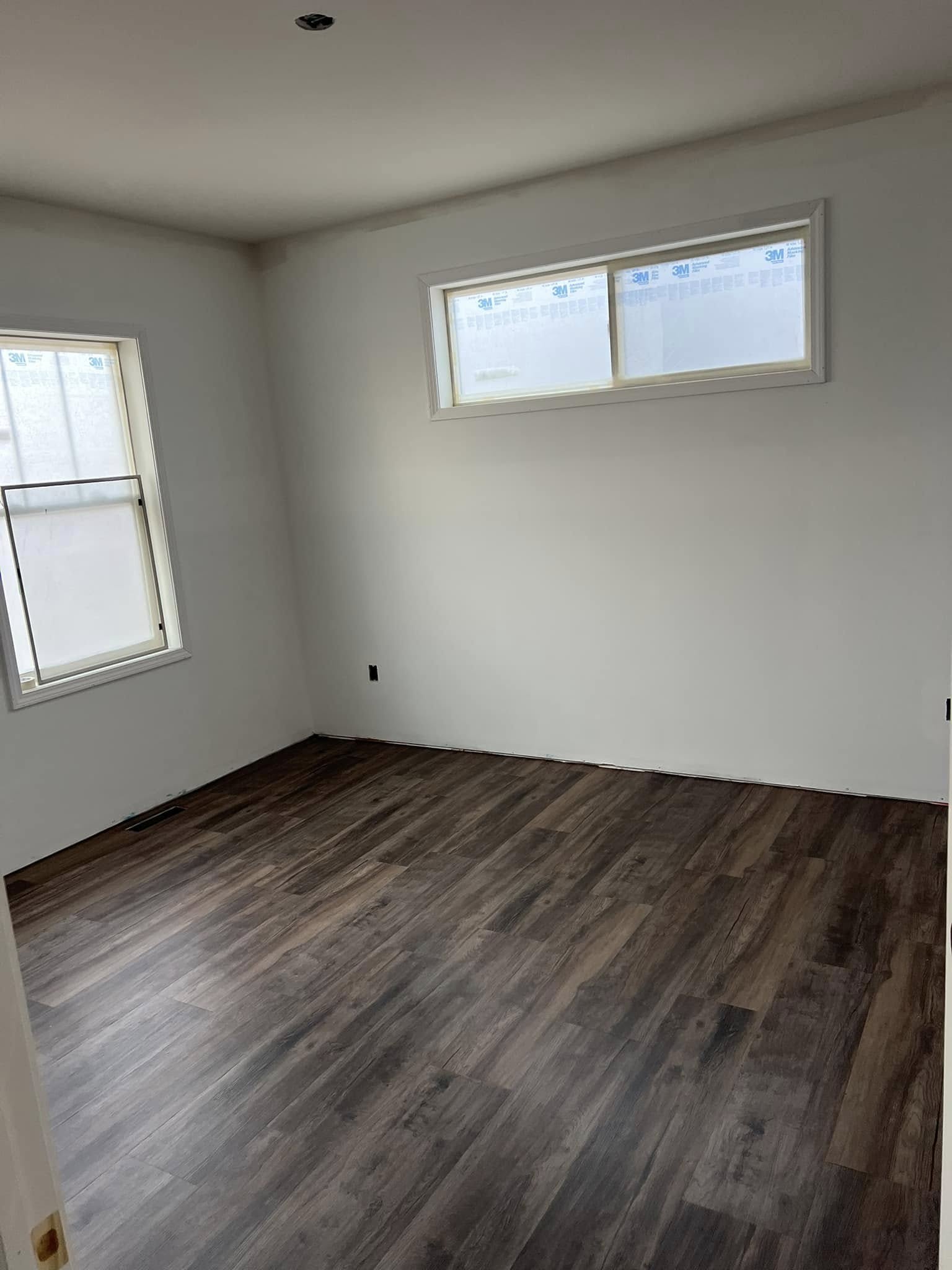 Empty room with light gray walls and dark wood flooring. Two windows allow natural light, creating a bright and calm atmosphere. Minimalist setting.