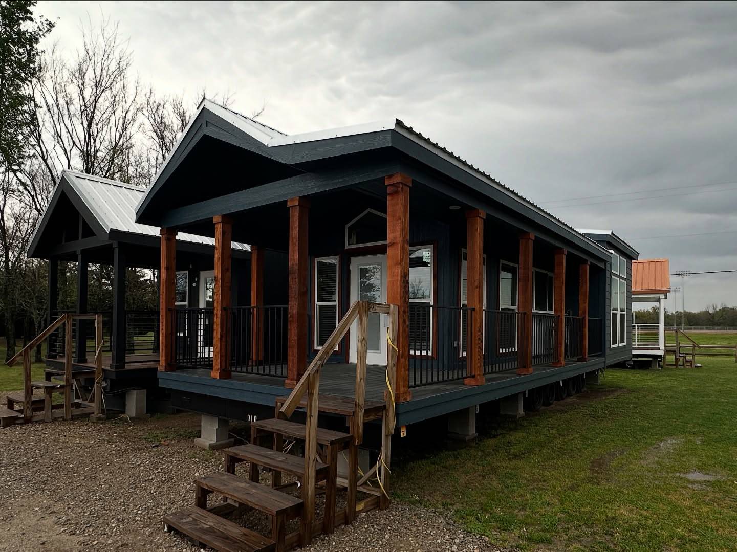 Alt text: "Three small, dark gray tiny houses with wooden pillars and metal roofs, set on green grassy land under a cloudy sky, displaying rustic charm."