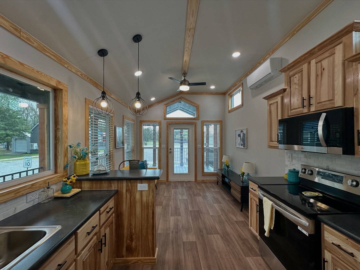 Modern kitchen with wooden cabinetry, black countertops, and stainless steel appliances. Pendant lights hang above, adding warmth. Large windows offer natural light.