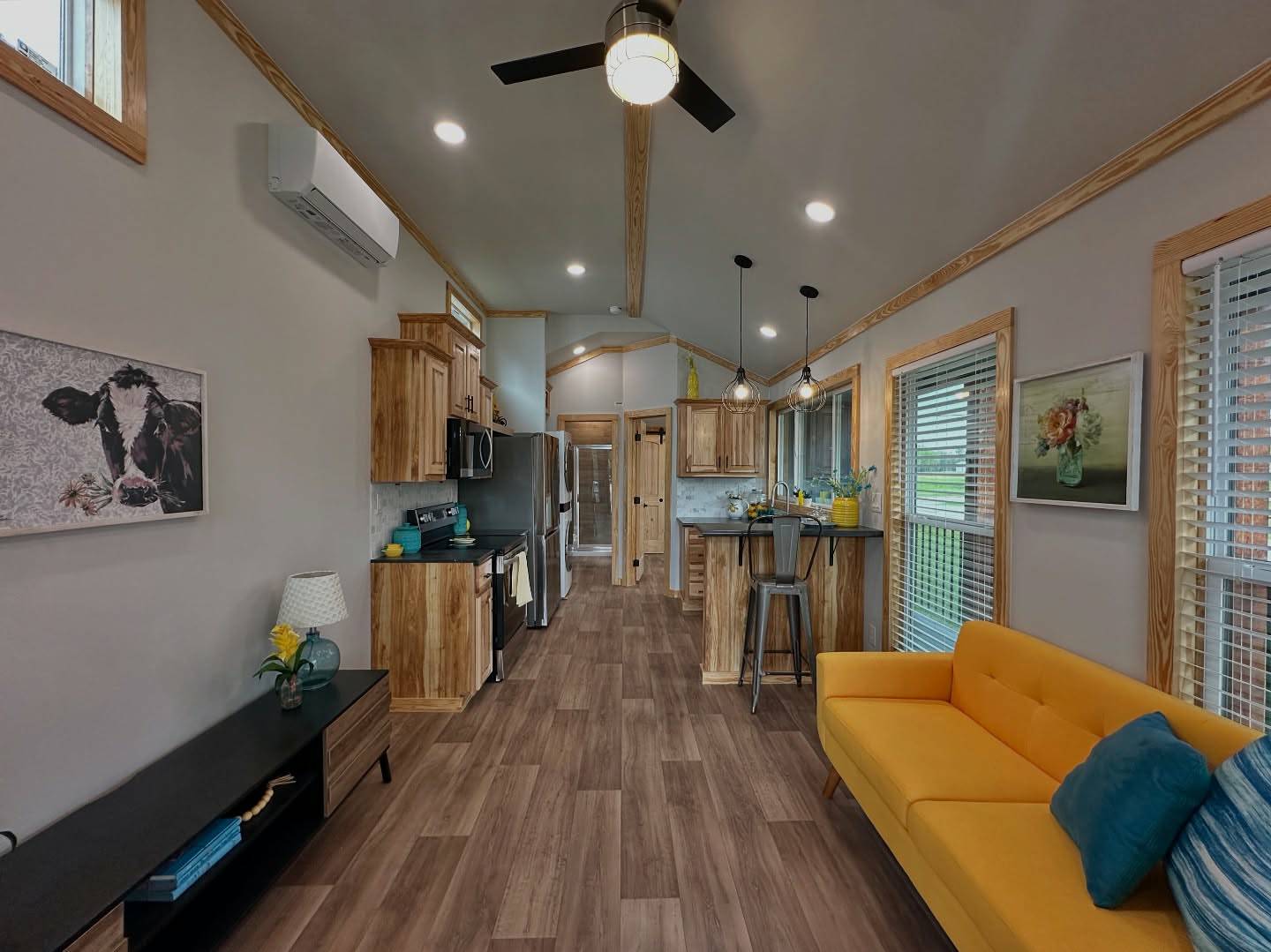 A stylish, modern tiny home interior with wood finishes, featuring a vibrant yellow couch, cow art, a sleek kitchen with an island, and pendant lighting.
