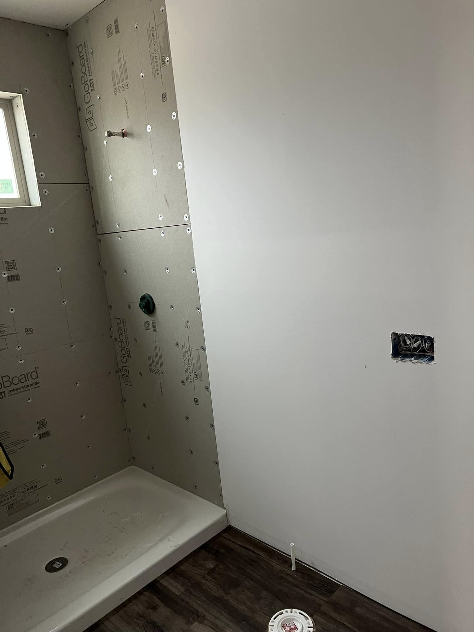 Partially renovated bathroom with drywall on walls, a shower base in the corner, and wooden flooring. A cutout for an electrical outlet is visible.