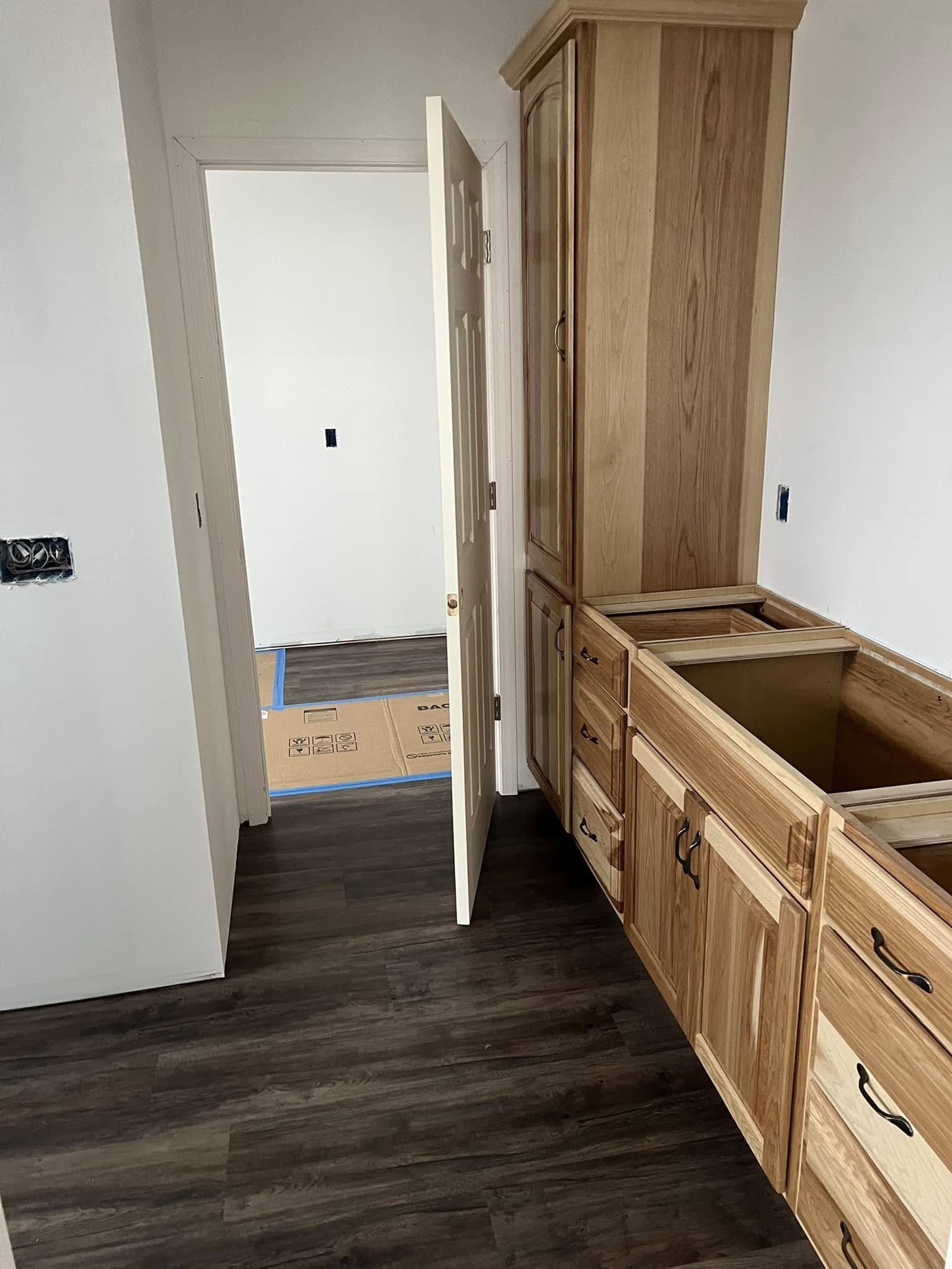 Wood cabinets line a narrow hallway with dark hardwood floors. A slightly open door reveals a room with a covered floor, suggesting ongoing renovation.
