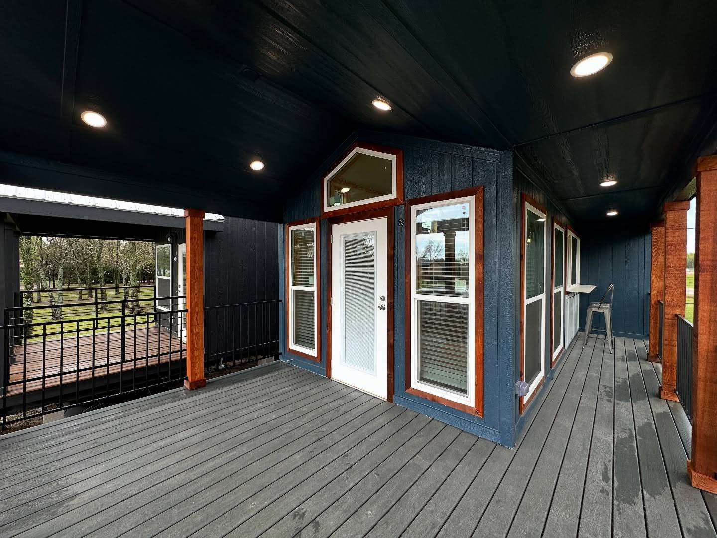 Covered patio with gray decking and recessed lighting. Blue walls and a white door with large windows offer a modern feel. Wooden posts add warmth.