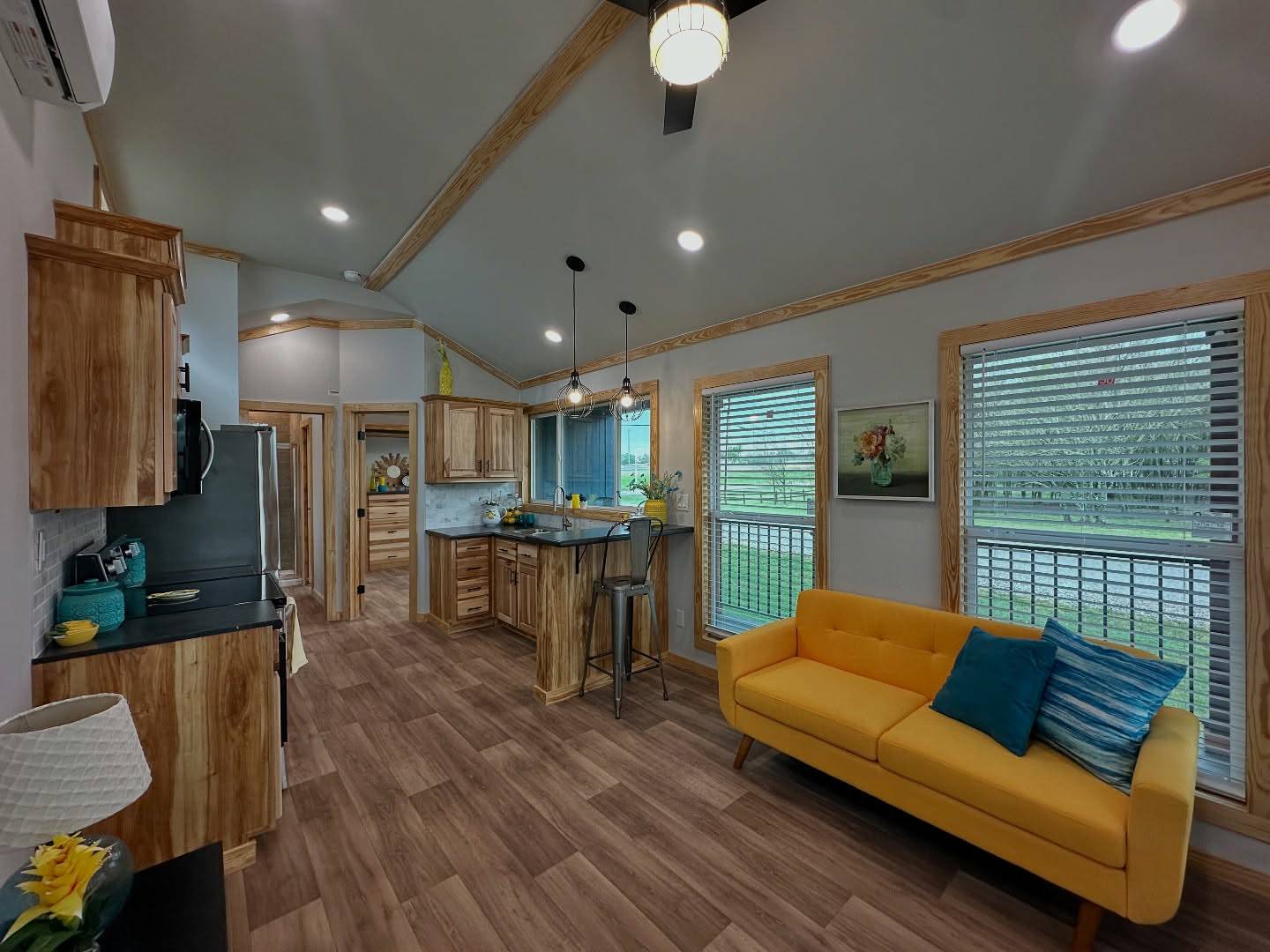 Modern living area with wood accents, featuring a bright yellow sofa with blue cushions. An open kitchen with light wood cabinets, pendant lights, and a breakfast bar. Large windows allow natural light, creating a warm and inviting space.