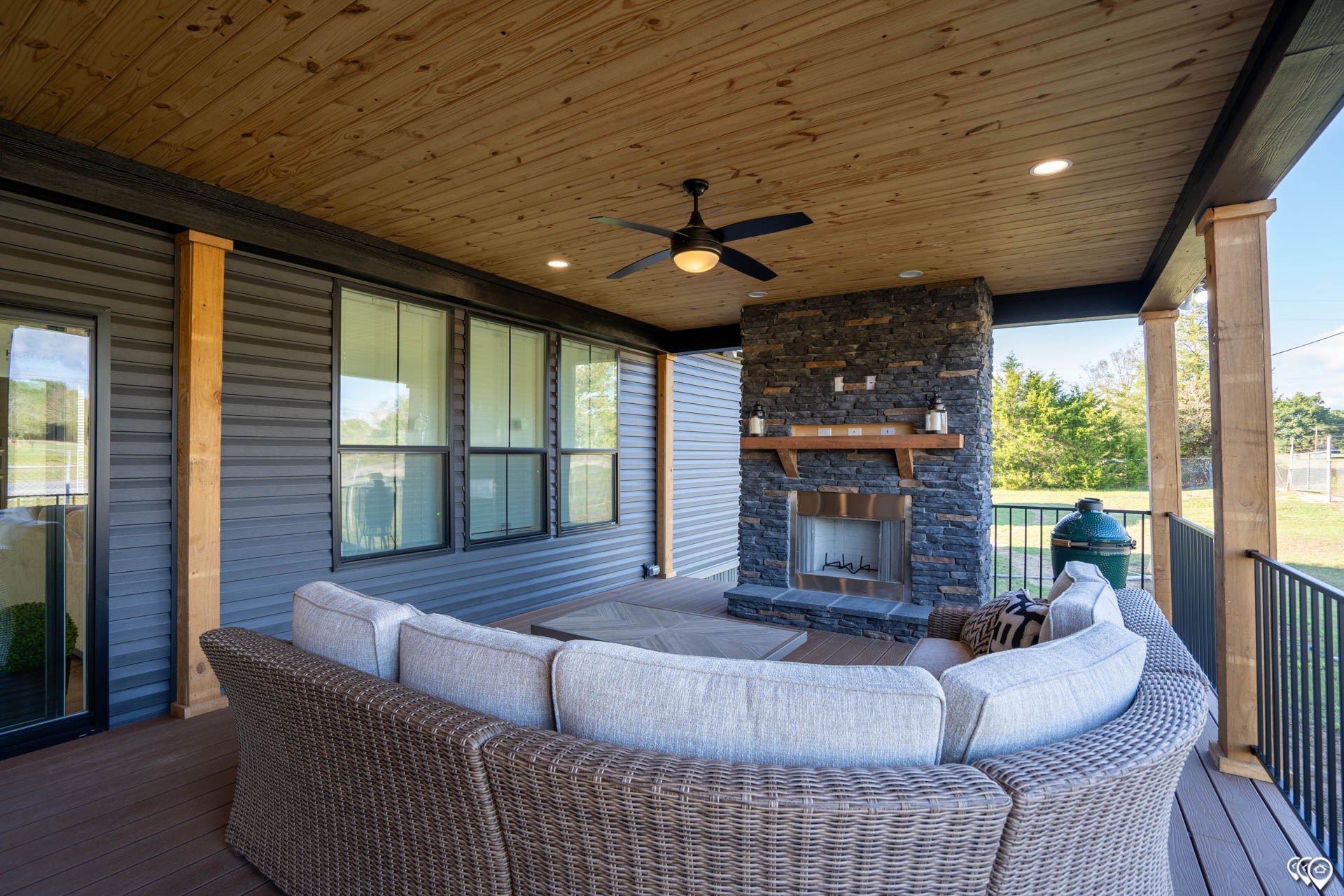 Covered patio with wicker sectional sofa facing a stone fireplace. Wood-paneled ceiling with a fan, cozy atmosphere, and a view of the backyard.
