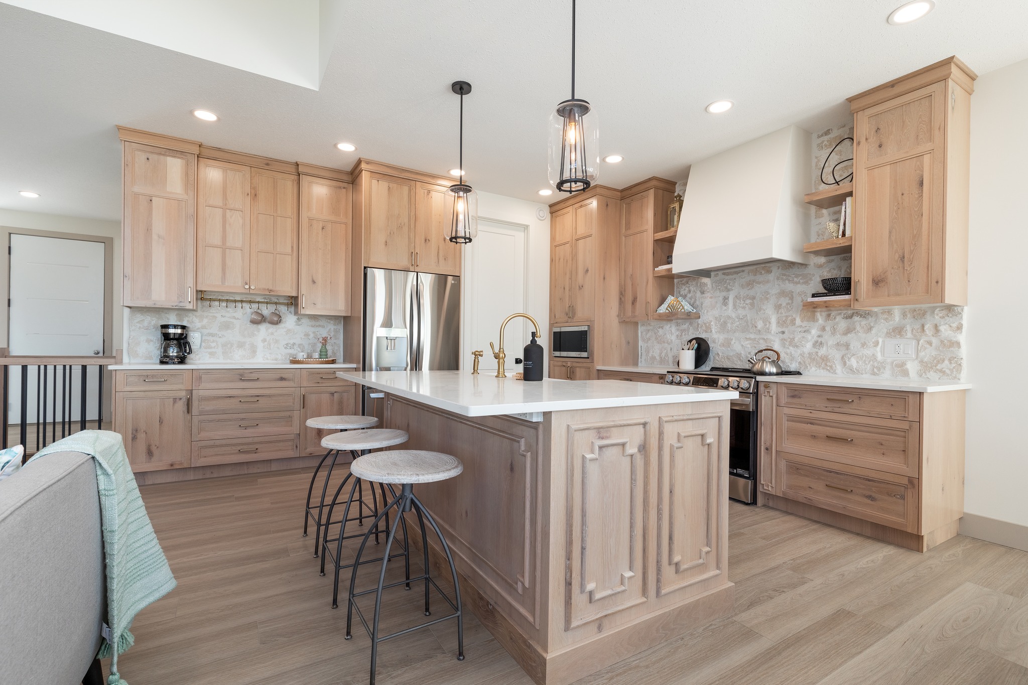 Elegant kitchen with light wood cabinets, a large island, and three barstools. Features stainless steel appliances and modern pendant lighting.