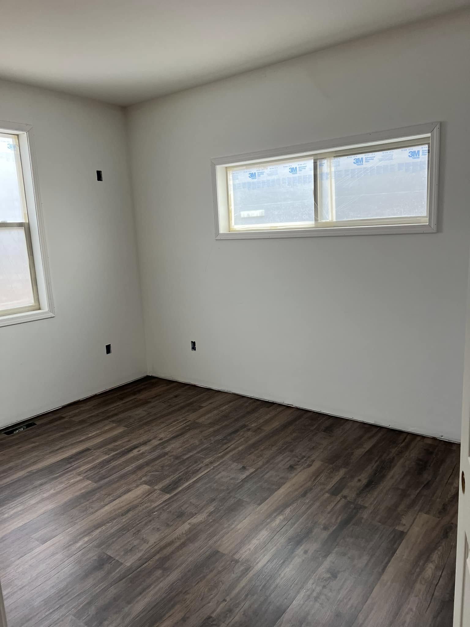 A minimalist, empty room with white walls and dark wooden flooring. Natural light streams through two rectangular windows, creating a calm and clean atmosphere.
