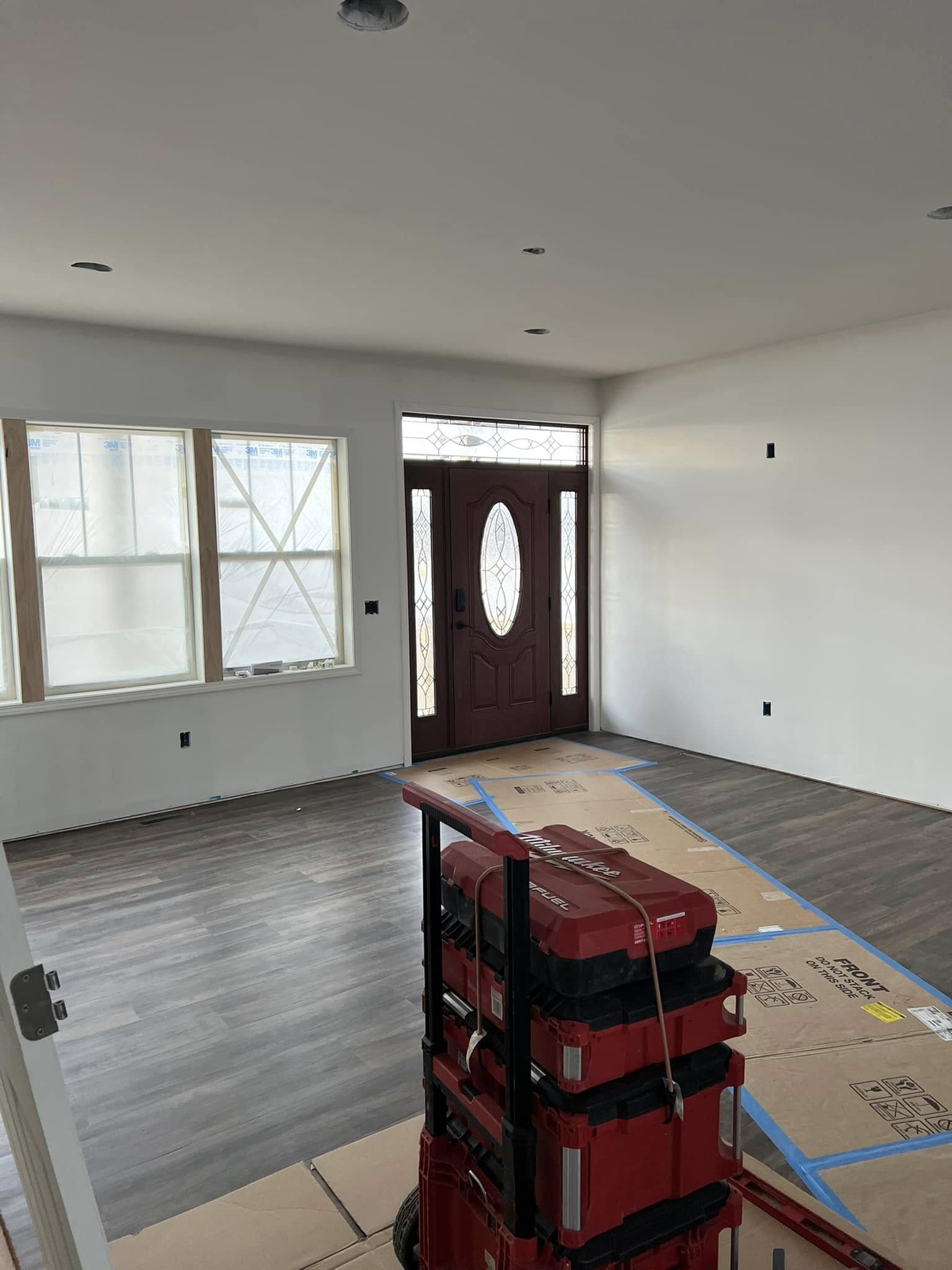 Unfurnished room with light gray walls and dark wood floors. A red door with glass panels is visible. Moving boxes and tools are in the foreground.