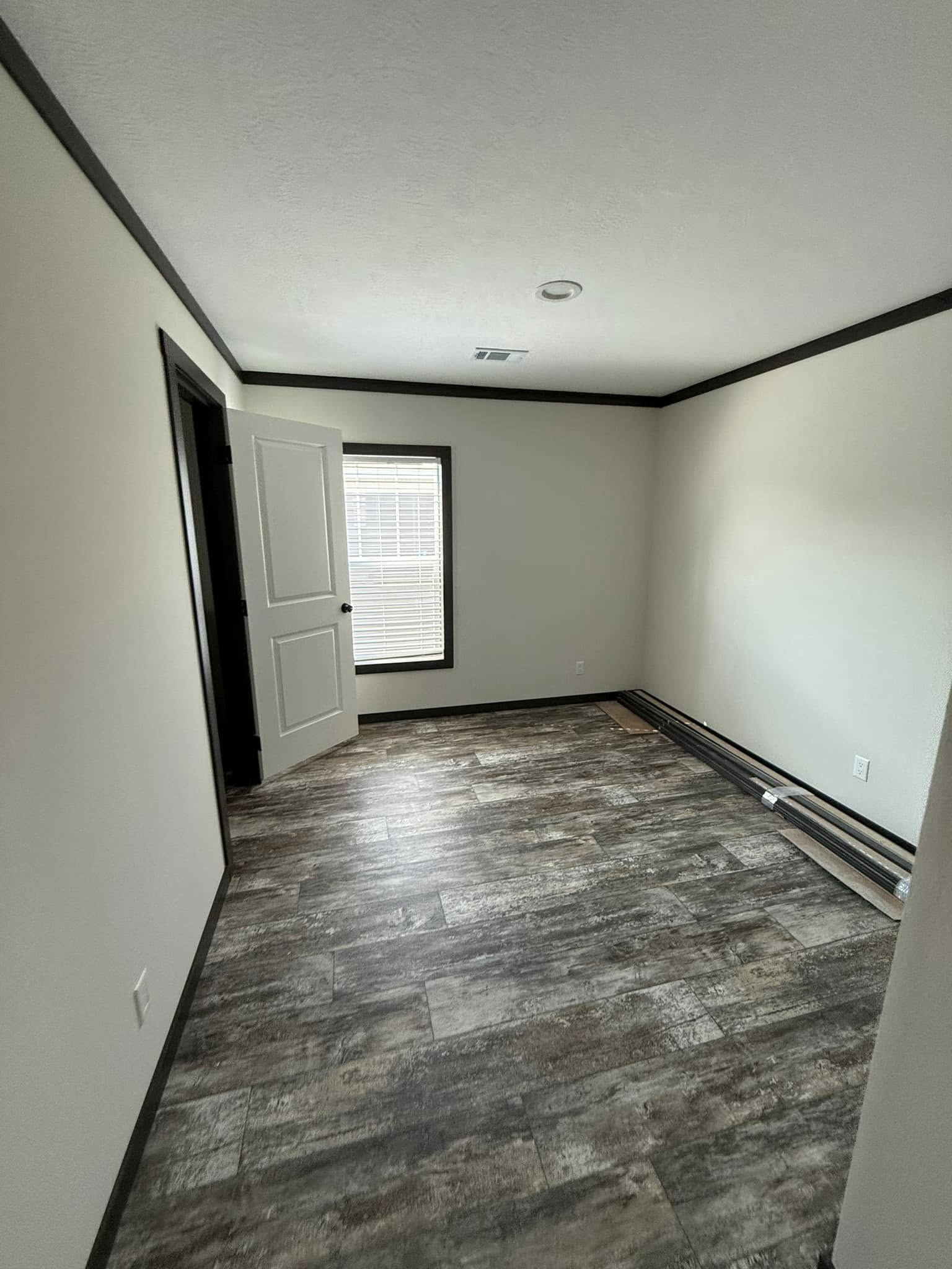 An empty room with beige walls, dark trim, and textured gray laminate flooring. A door is ajar near a window with blinds, offering soft natural light.