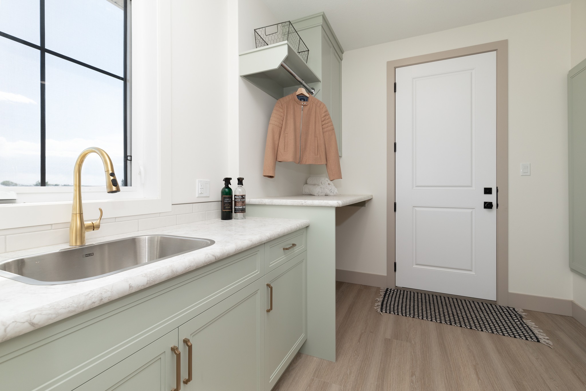 A minimalist laundry room features light green cabinets, a marble countertop with a gold faucet, and a doorway. A tan coat hangs under a cabinet.