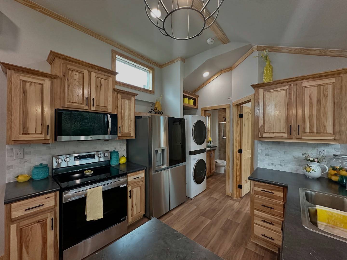 Compact kitchen with wooden cabinets, stainless steel appliances, and a stacked washer and dryer. Bright natural light, organized, cozy, modern ambiance.