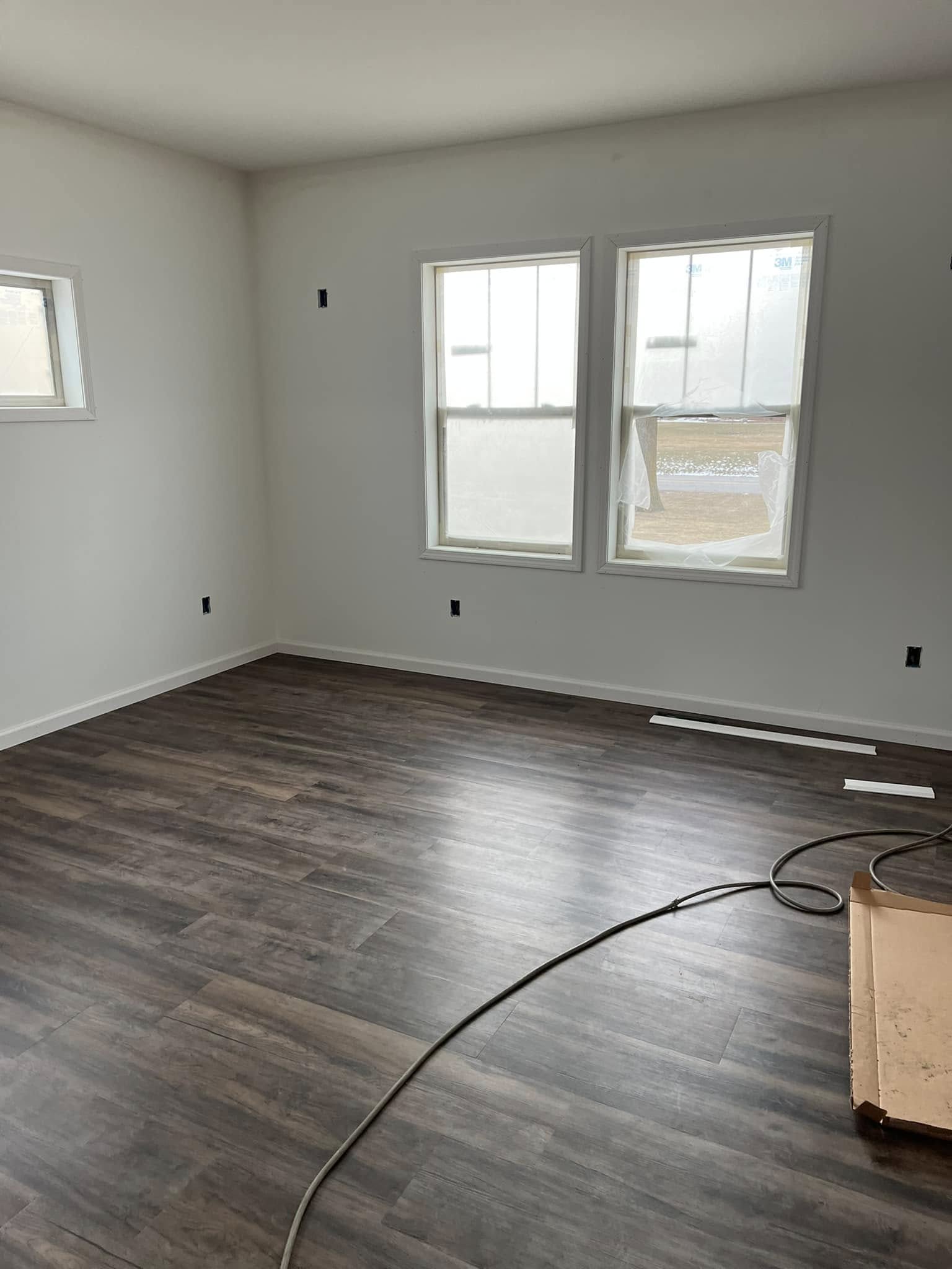 A freshly renovated room with white walls and dark wood flooring. Two large windows let in natural light, and a cable and cardboard box rest on the floor.