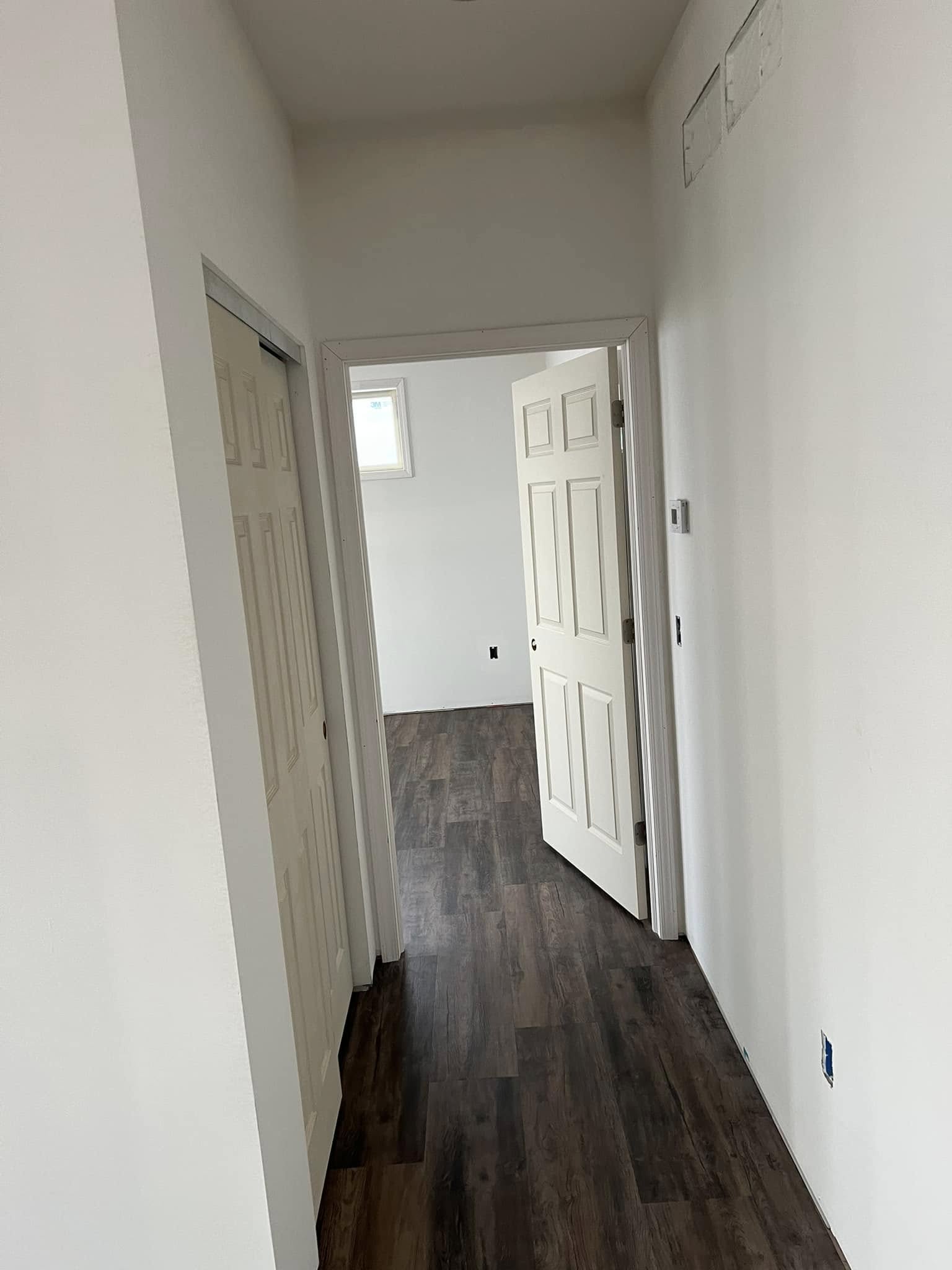 Narrow hallway with white walls and dark wooden flooring. Closed closet door on the left and an open door at the end, revealing a sunlit room. Serene atmosphere.