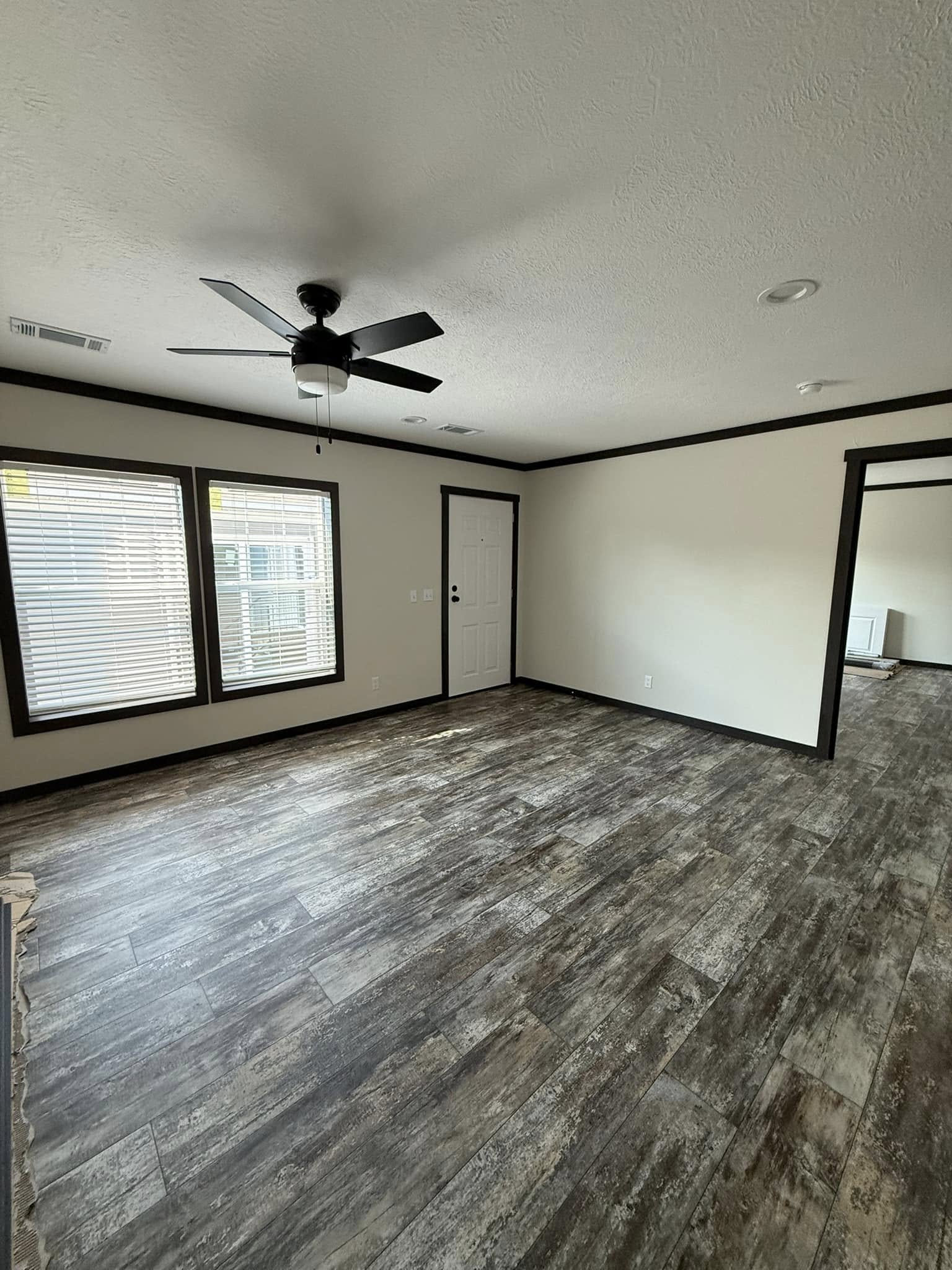Empty room with dark wood flooring and light gray walls. A ceiling fan is centered on the white ceiling. Two large windows emit natural light next to a white door.