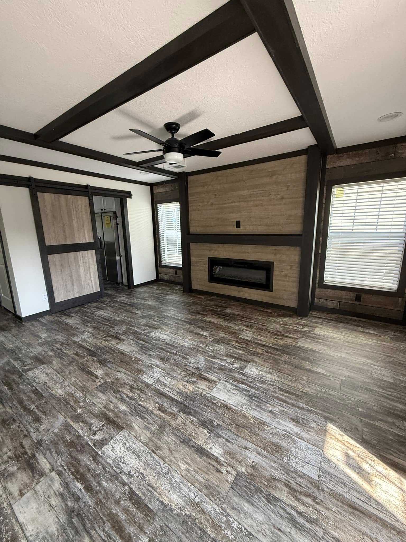 Spacious room with rustic wood flooring and beams, featuring a sleek electric fireplace. Two large windows allow natural light, enhancing the warm, cozy atmosphere.