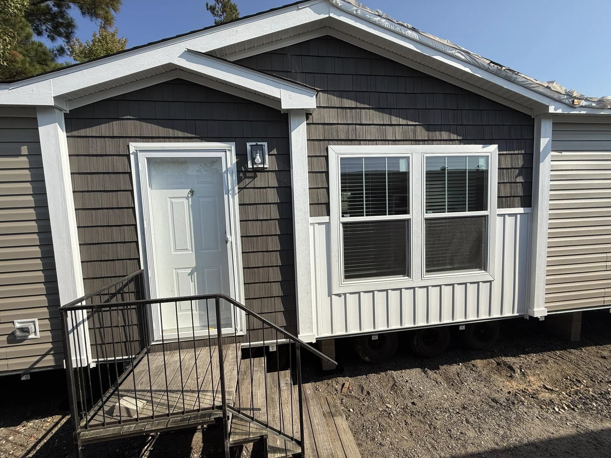 A compact, gray mobile home with white accents, featuring a closed white door, two windows, and a small wooden porch with a metal railing. Bright, clear day.