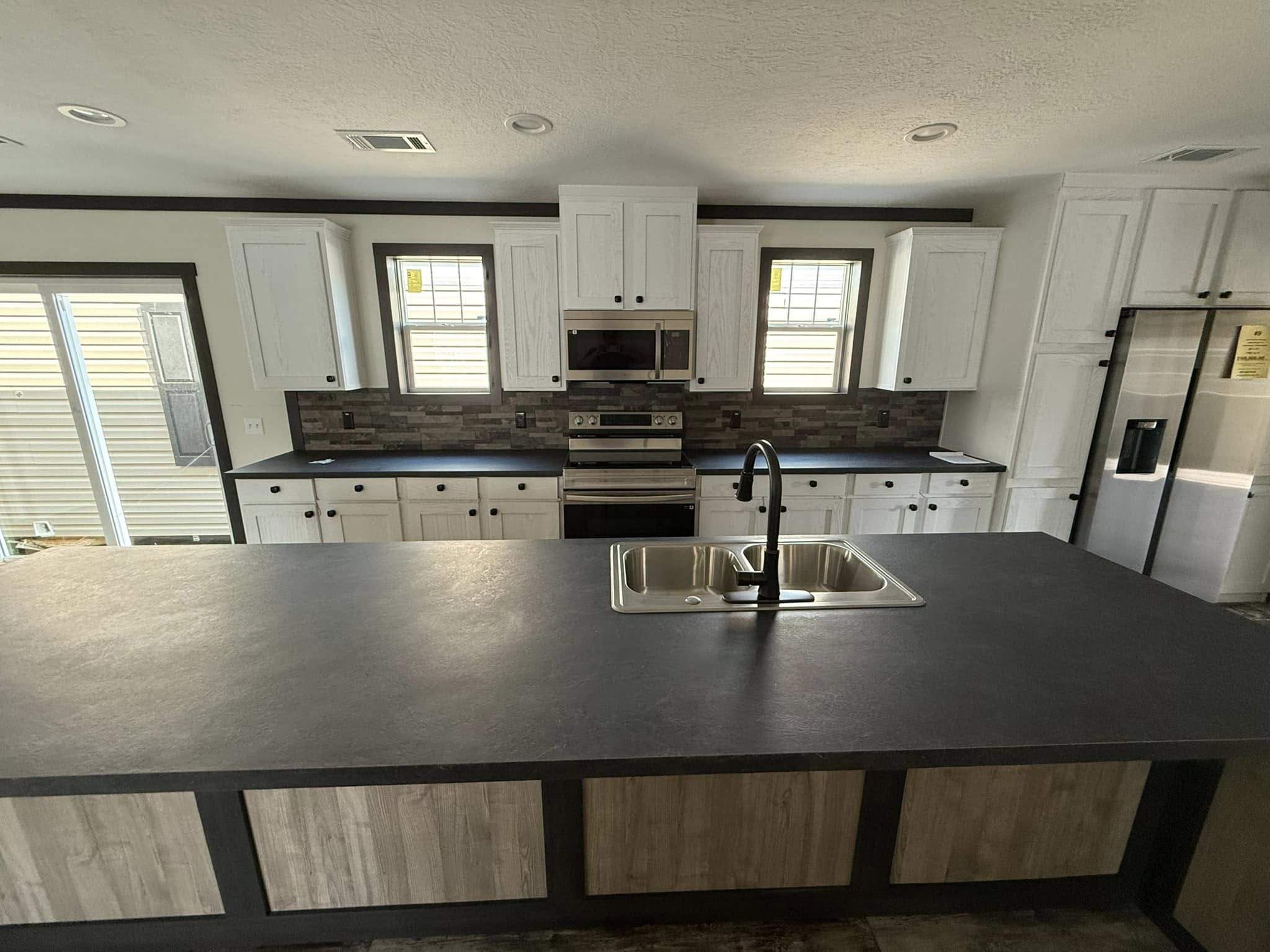 Modern kitchen with large island, double sink, dark countertop, and light wood cabinets. Stainless steel appliances and backsplash create a sleek feel.