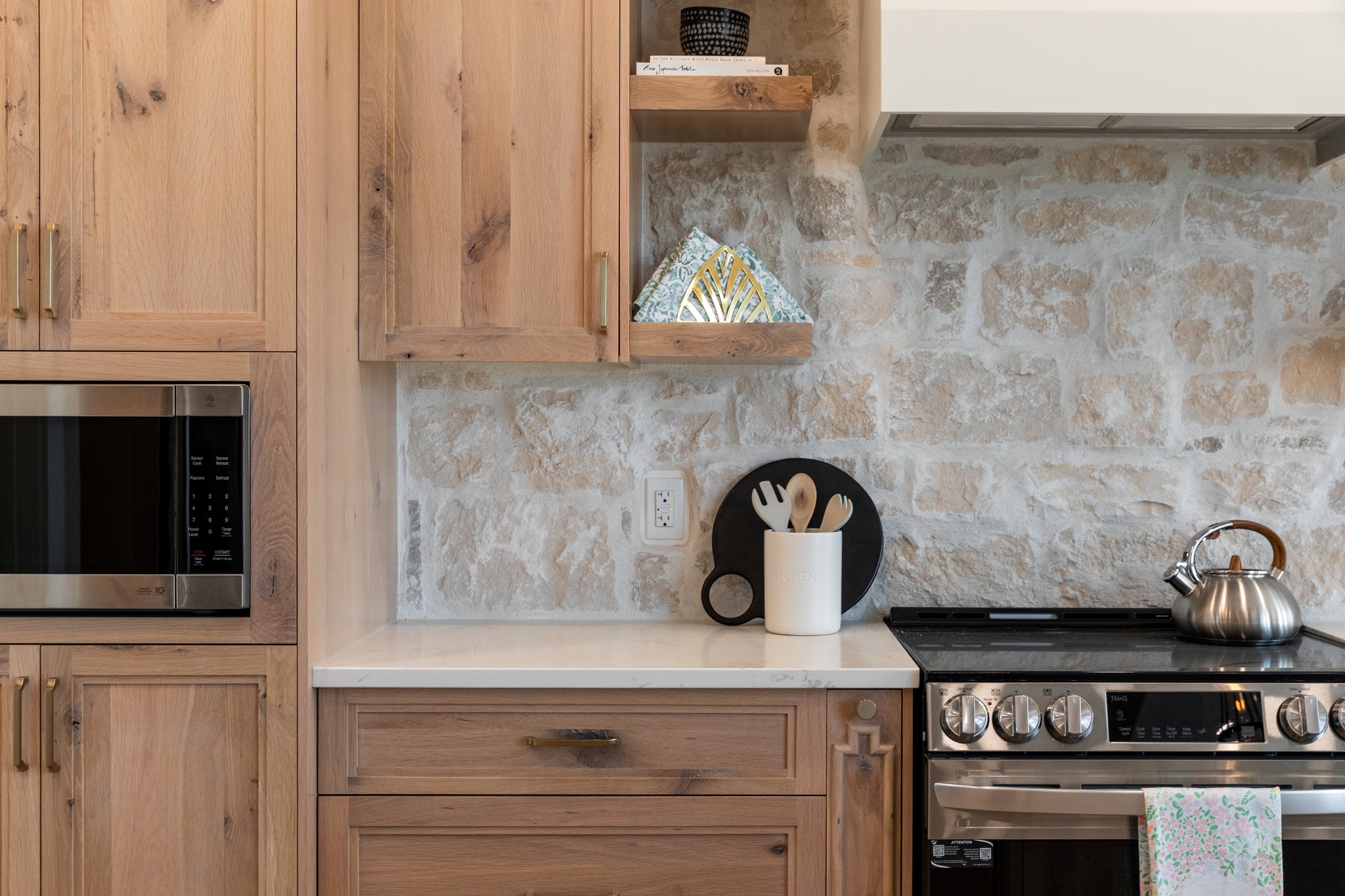 Modern kitchen with warm wooden cabinets and stone backsplash. Features a microwave, utensils in a holder, and a stainless steel kettle on the stove. Cozy and inviting.
