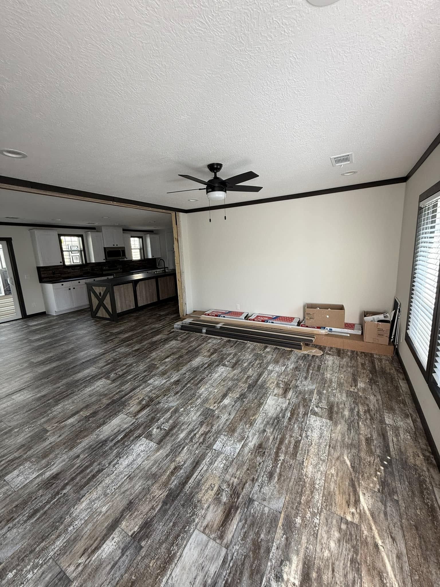 Spacious living room with rustic wood flooring, an overhead ceiling fan, and large windows. Renovation materials are stacked against a light-colored wall.