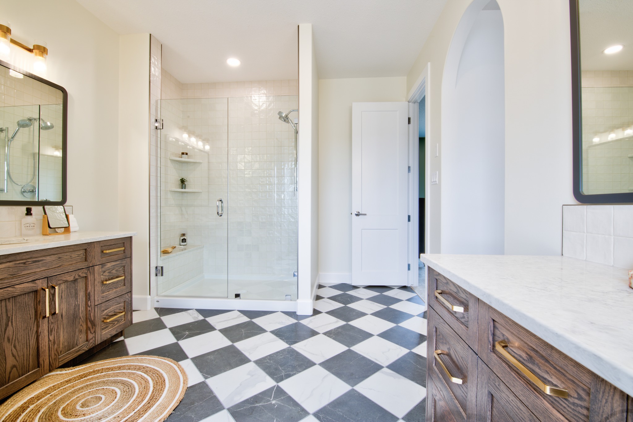 Spacious bathroom with two wood vanities and a large glass shower. Checkered floor, modern lighting, and white walls create an elegant, inviting atmosphere.
