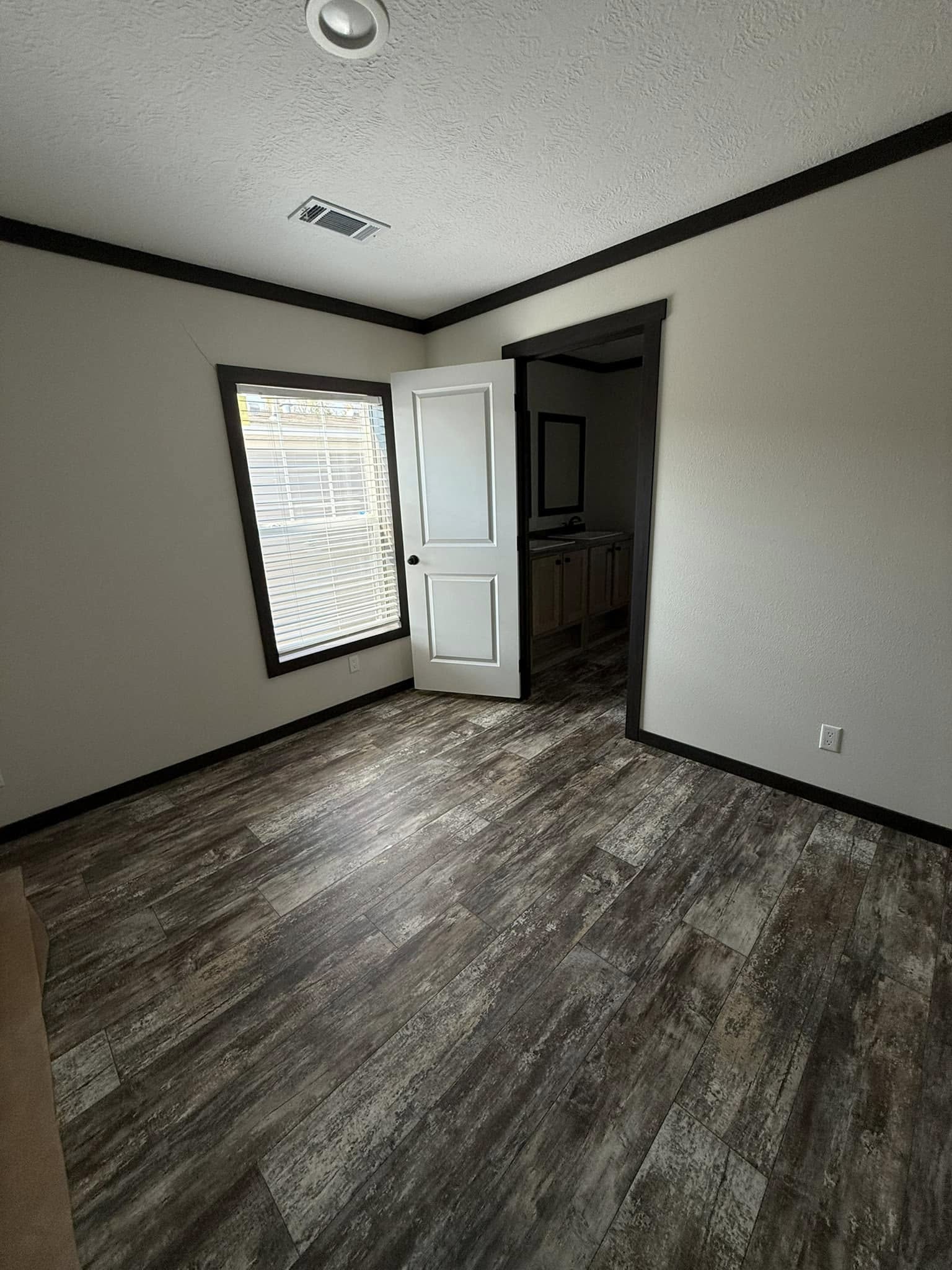 Empty room with wood-patterned flooring and light-colored walls. A window with blinds allows natural light. An open door leads to another area.