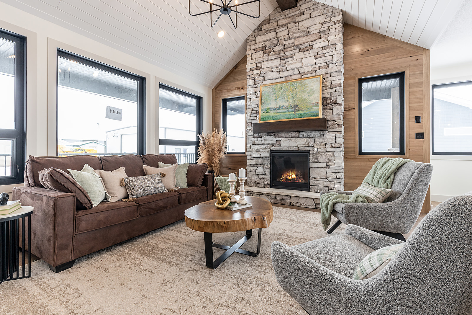 Stylish living room with stone fireplace, brown sofa, and gray chairs. Warm tones, large windows, abstract art, and cozy, inviting atmosphere.