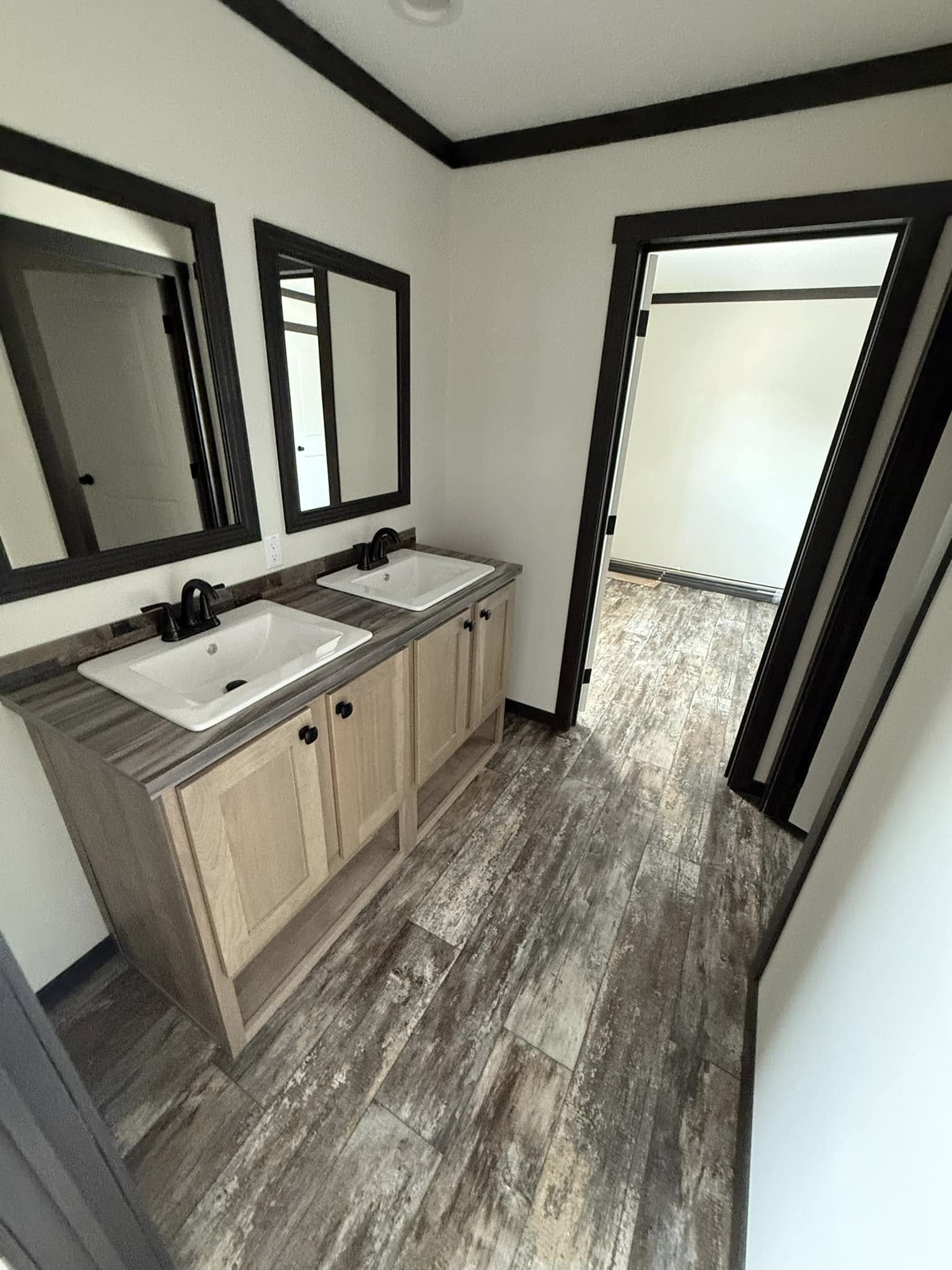 A modern bathroom features a brown wood floor, double sinks with black faucets, and black-framed mirrors. Neutral tones create a clean, spacious feel.