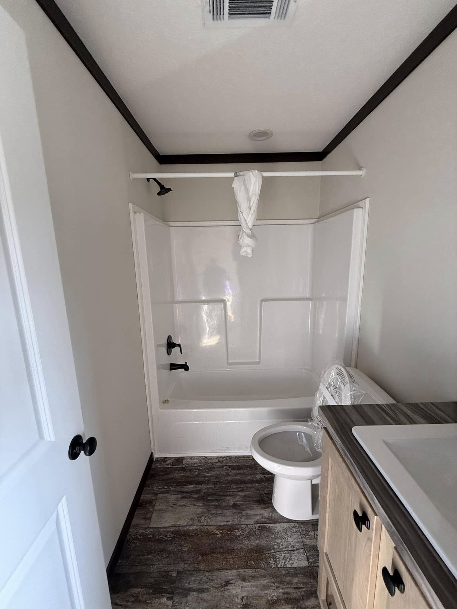 Compact bathroom with white shower-tub combo, dark fixtures, and wooden cabinetry. Towel is hanging atop the shower rod, creating a tidy, minimalistic feel.