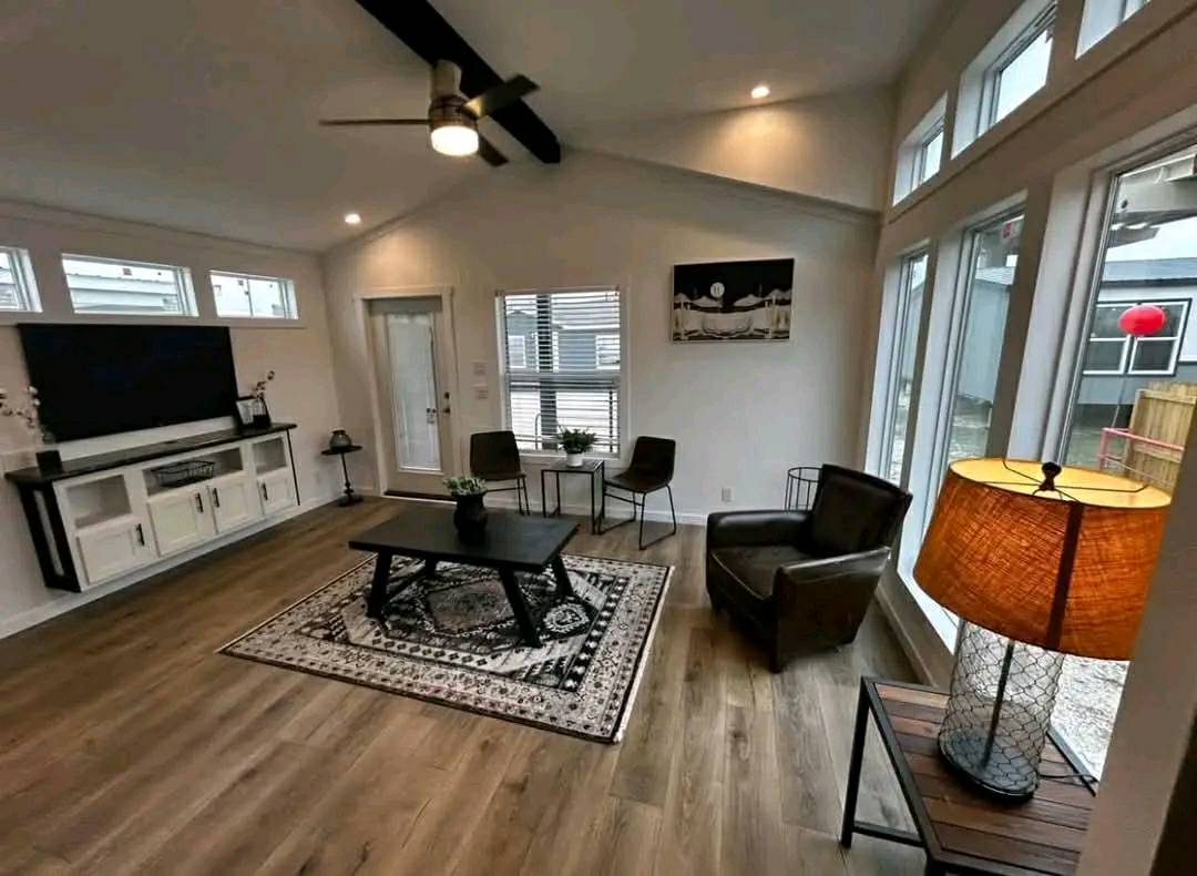 Spacious modern living room with wood flooring, large windows, and a black TV on a white stand. Brown chairs, a patterned rug, and soft lighting create a cozy ambiance.