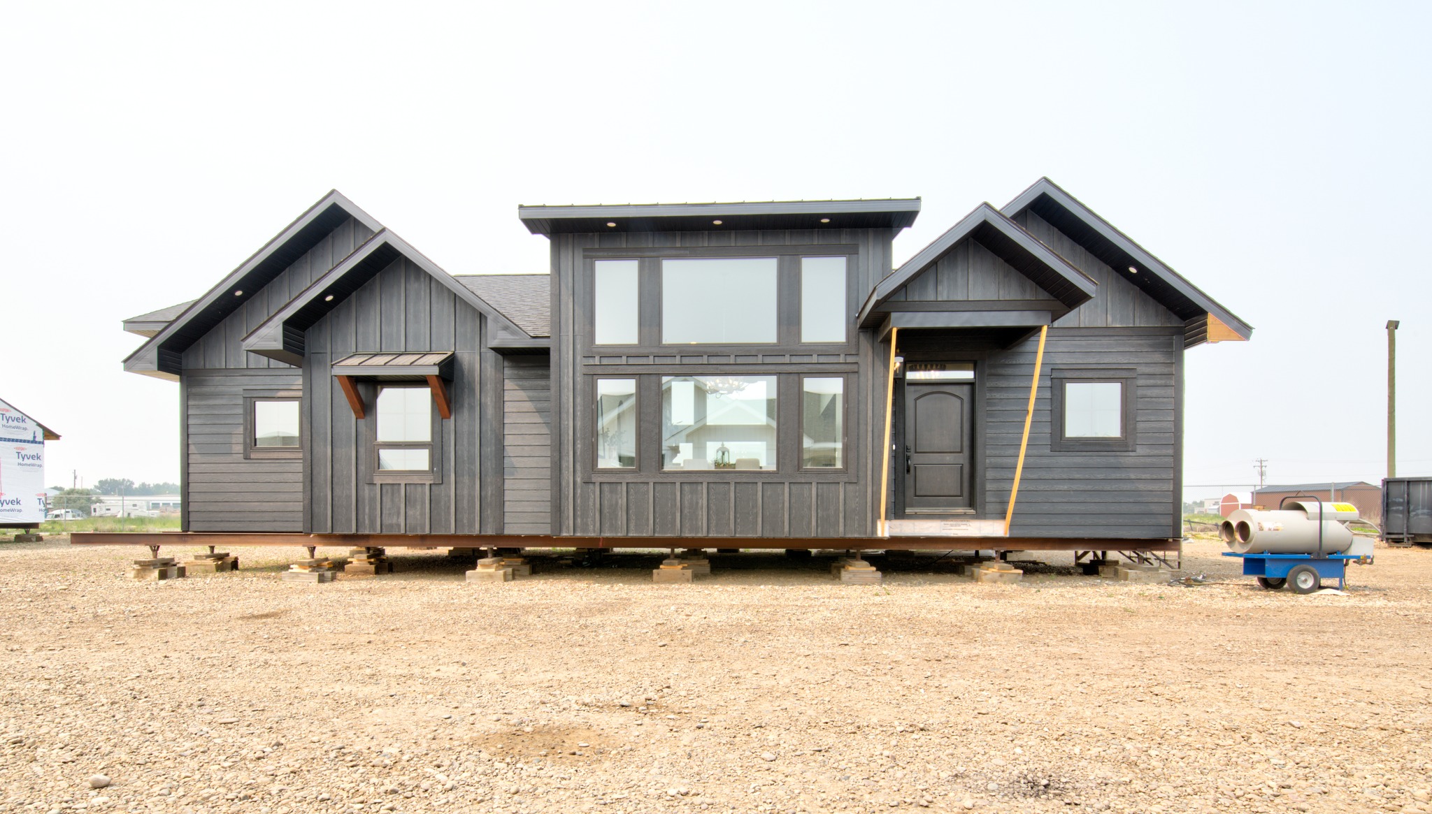 A gray prefabricated house stands on wooden blocks in a gravel lot. It features a modern design with large windows and a black door, evoking simplicity.