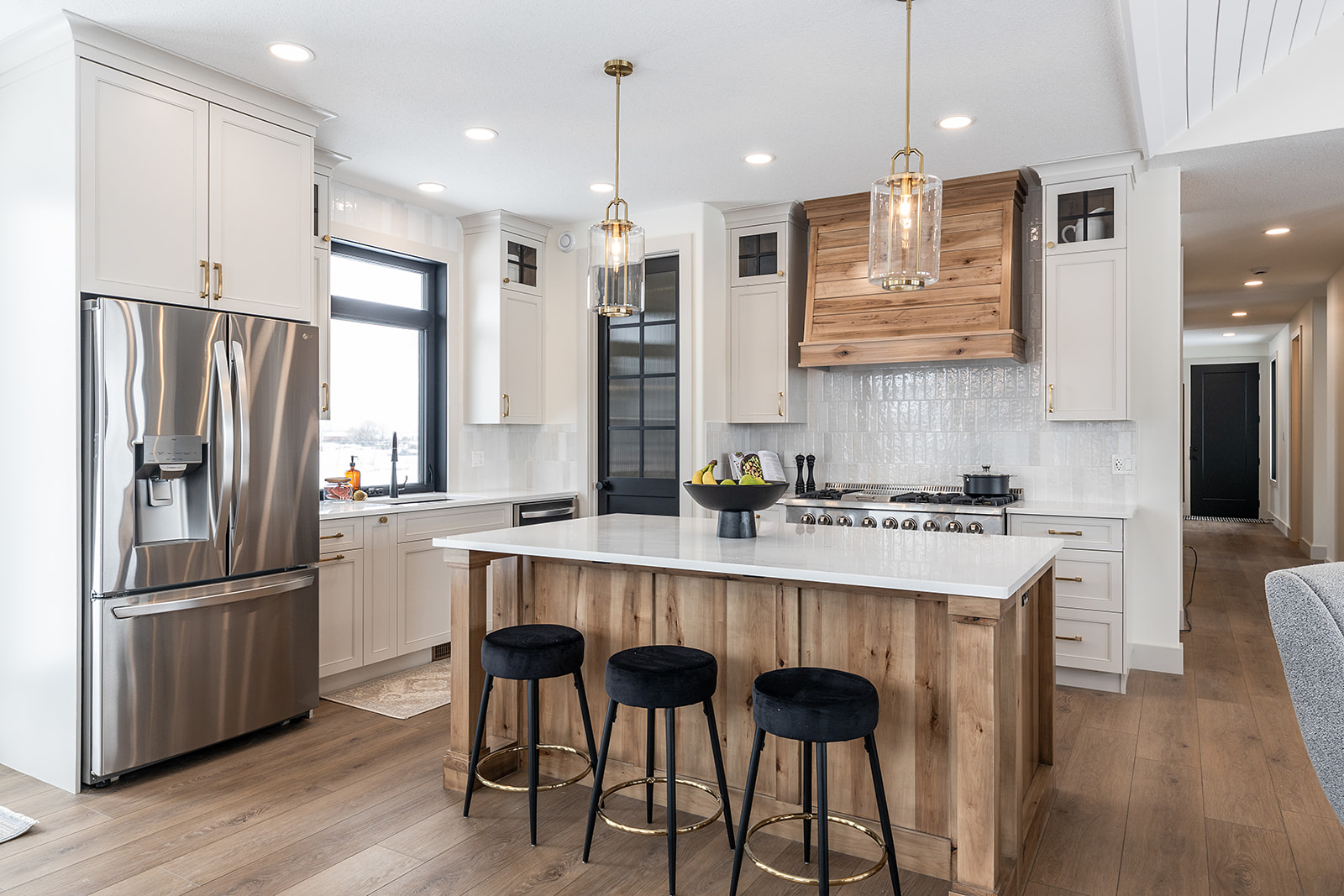 Modern kitchen with white cabinets, wooden island, and marble countertop. Three black stools, stainless steel fridge, and pendant lights enhance the sleek design.