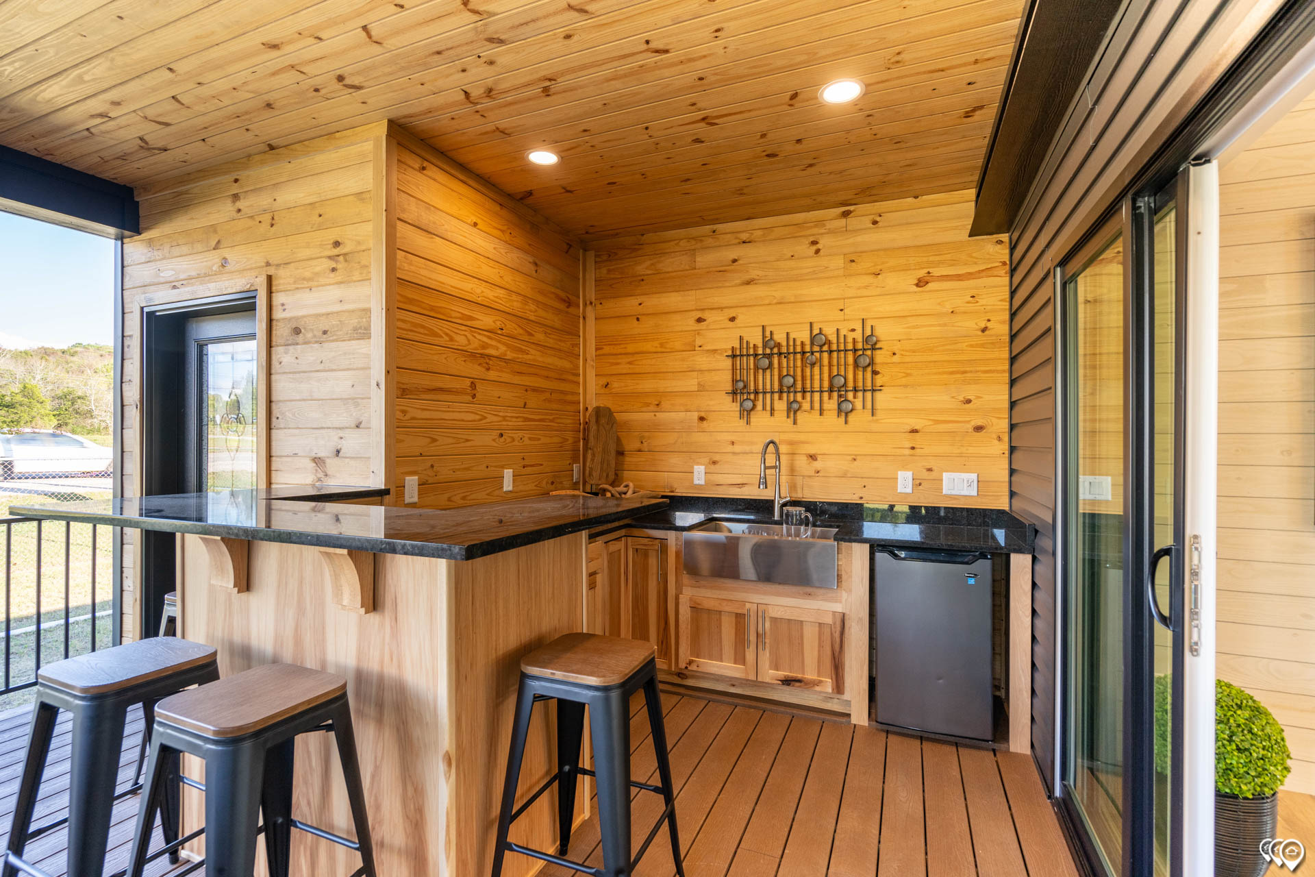 Cozy outdoor bar with wooden walls and ceiling, featuring a black countertop, three stools, small sink, fridge, and decorative wall art. Natural light filters in.