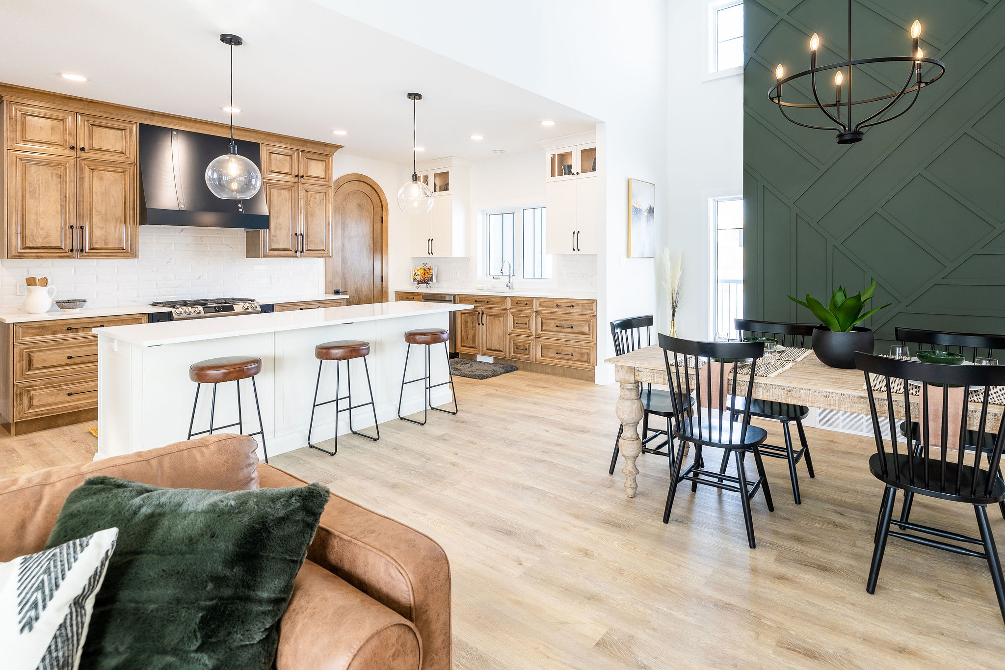A spacious modern kitchen and dining area with wooden cabinets, a large white island with three stools, pendant lights, and a dining table with black chairs.