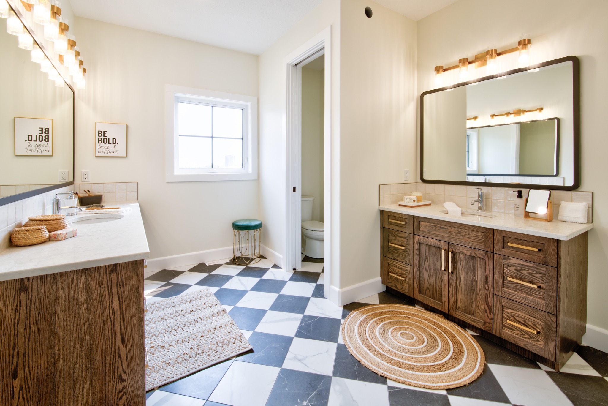 Spacious bathroom with checkered floor, dual wooden vanities, round rugs, and bright lighting. A window and framed quotes enhance the cozy ambiance.